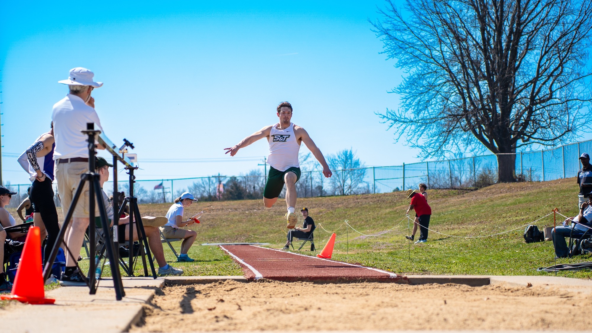 issouri S&T triple jumper Justin Chadwell soars through the air during an attempt at the Miner Invitational, approaching the sand pit as officials and spectators watch from the runway.