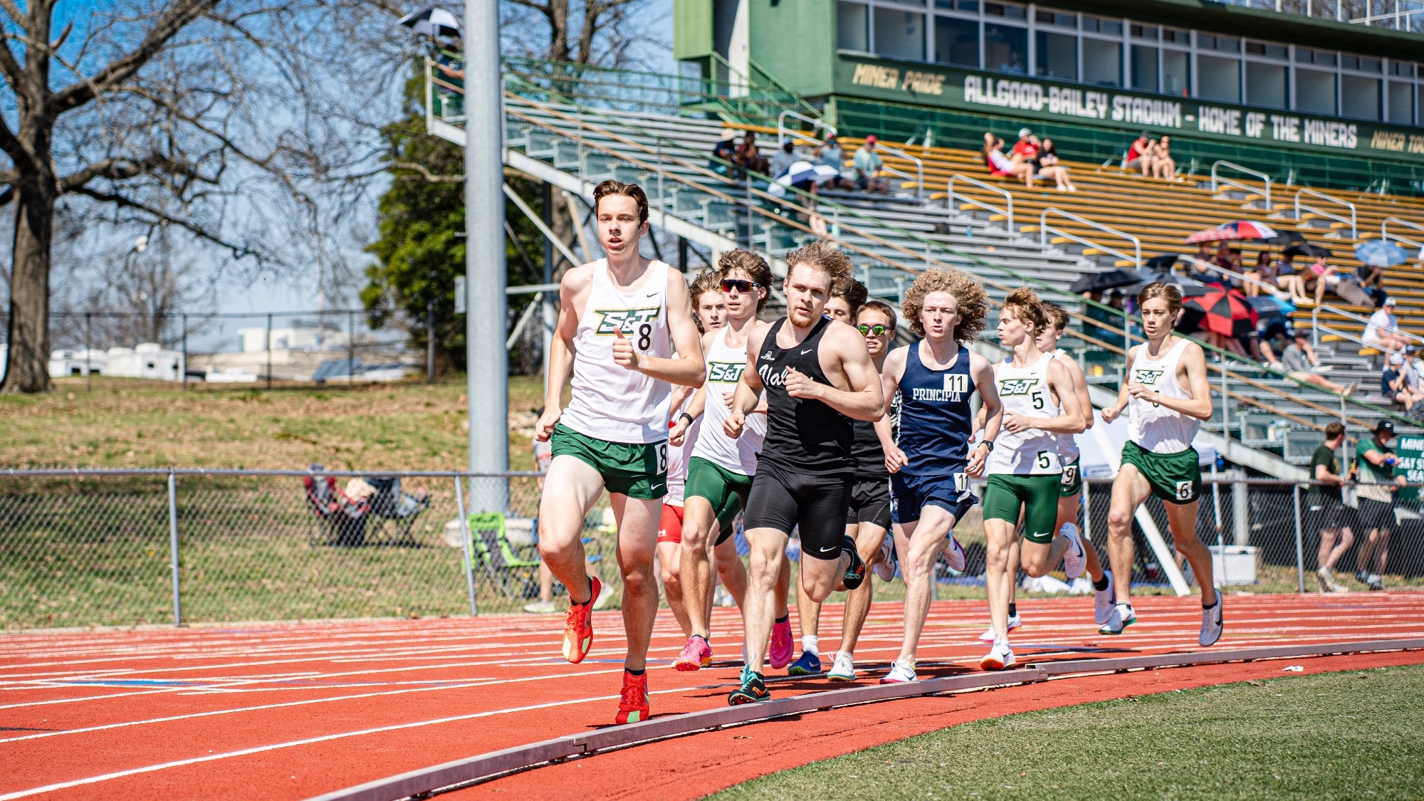 Missouri S&T distance runner Braden Stillmaker leads a pack of runners around the track during a race at the Miner Invitational at Allgood-Bailey Stadium, with competitors close behind and spectators in the stands.