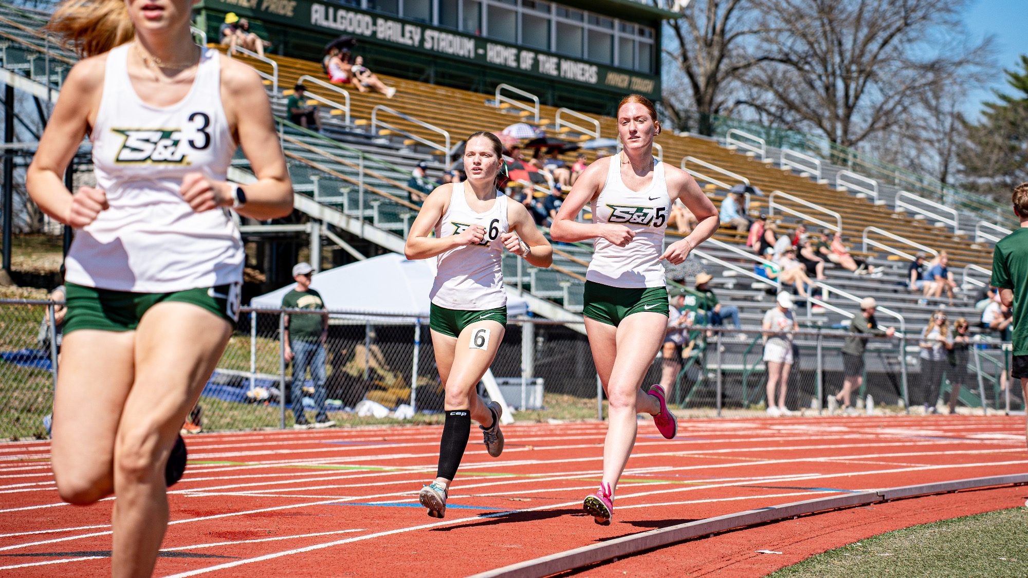 Missouri S&T distance runners Isabell Hacker (right) and Myra Roth (left) run side by side on the track during a race at Allgood-Bailey Stadium, with teammates and spectators in the background.