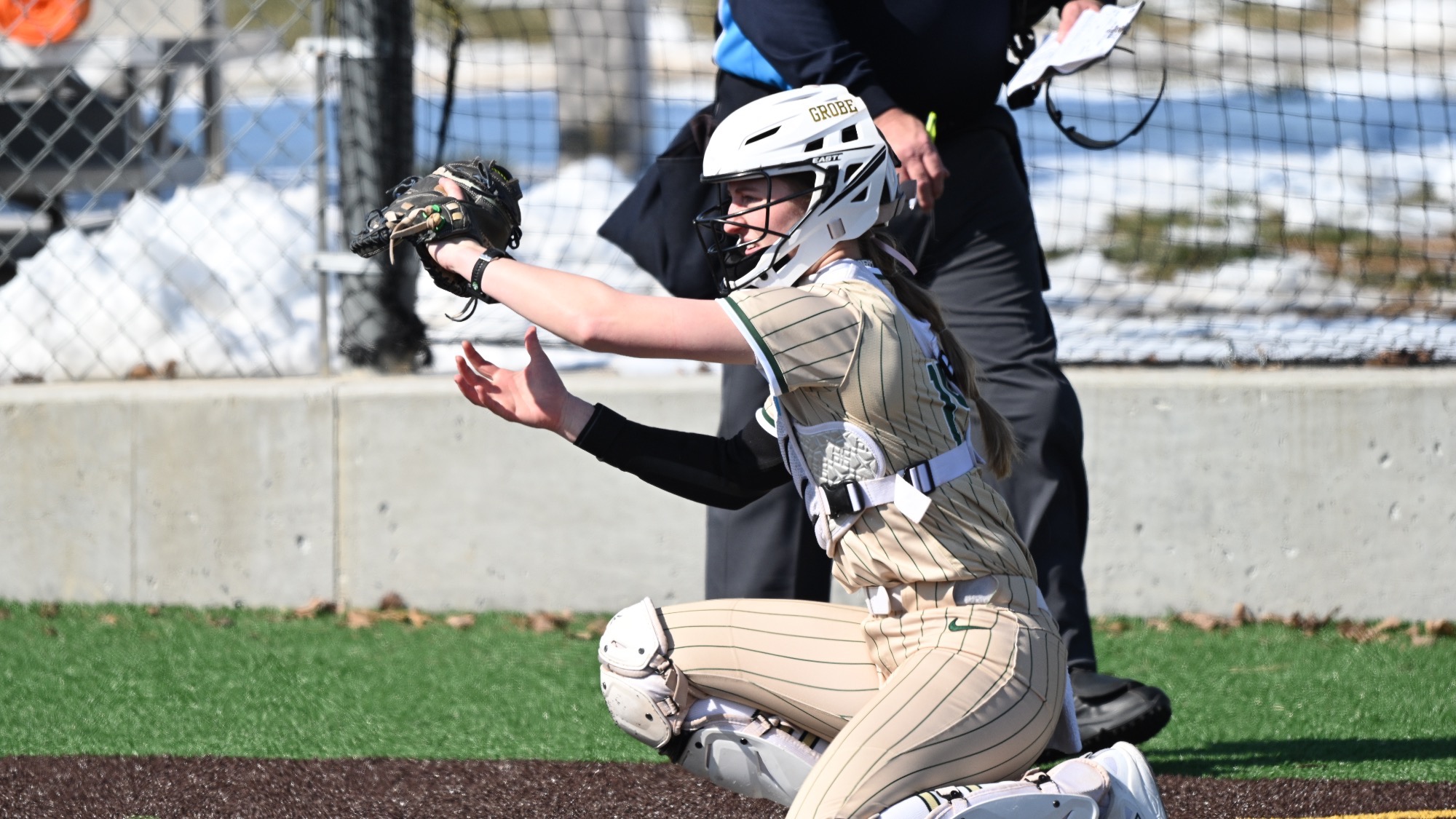 Missouri S&T catcher Trista Hunt, in full gear and a gold pinstripe jersey, receives a pitch while kneeling behind home plate as an umpire walks in the background during a softball game.