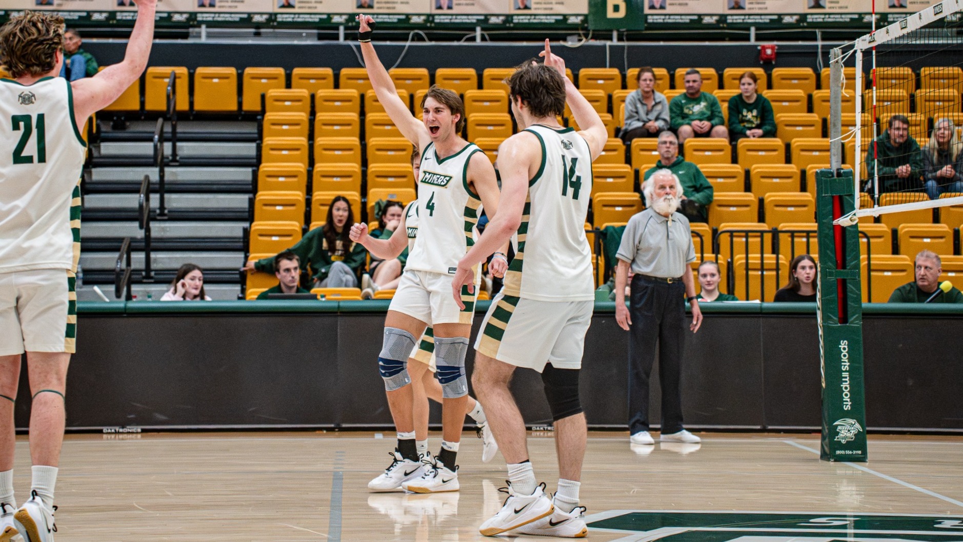 Tyler Johnwick, wearing number 4 in a white Missouri S&T volleyball uniform with green accents, raises his arm and celebrates a point near the net with teammate Aaron Sallade, number 14, during a match inside Gibson Arena. A third teammate joins the celebration at left while spectators sit in the yellow bleachers and player banners hang along the wall in the background.