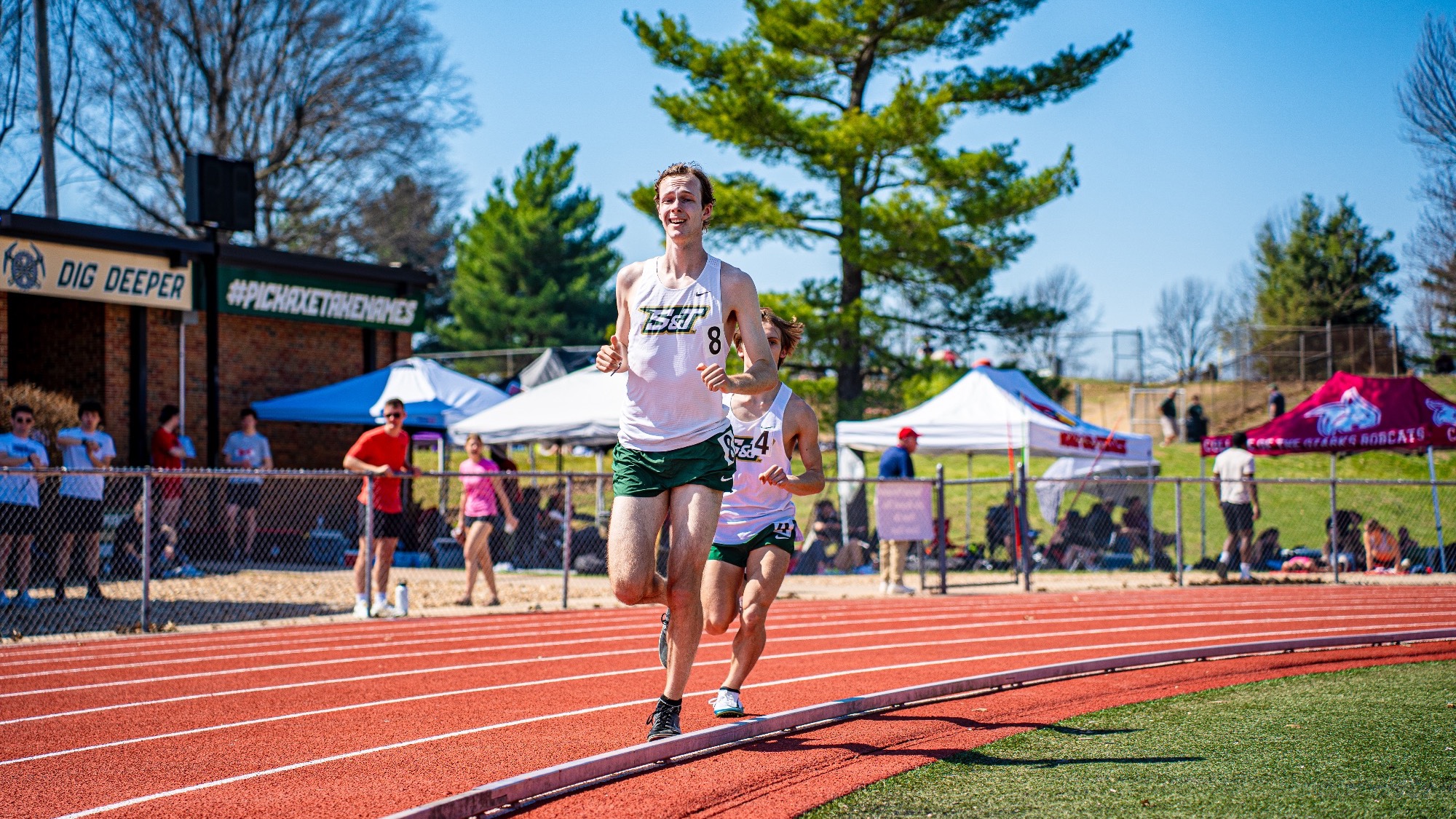Missouri S&T distance runner Maddox Riley leads down the track during a race at the Miner Invitational, with a teammate close behind and spectators lining the fence in the background.