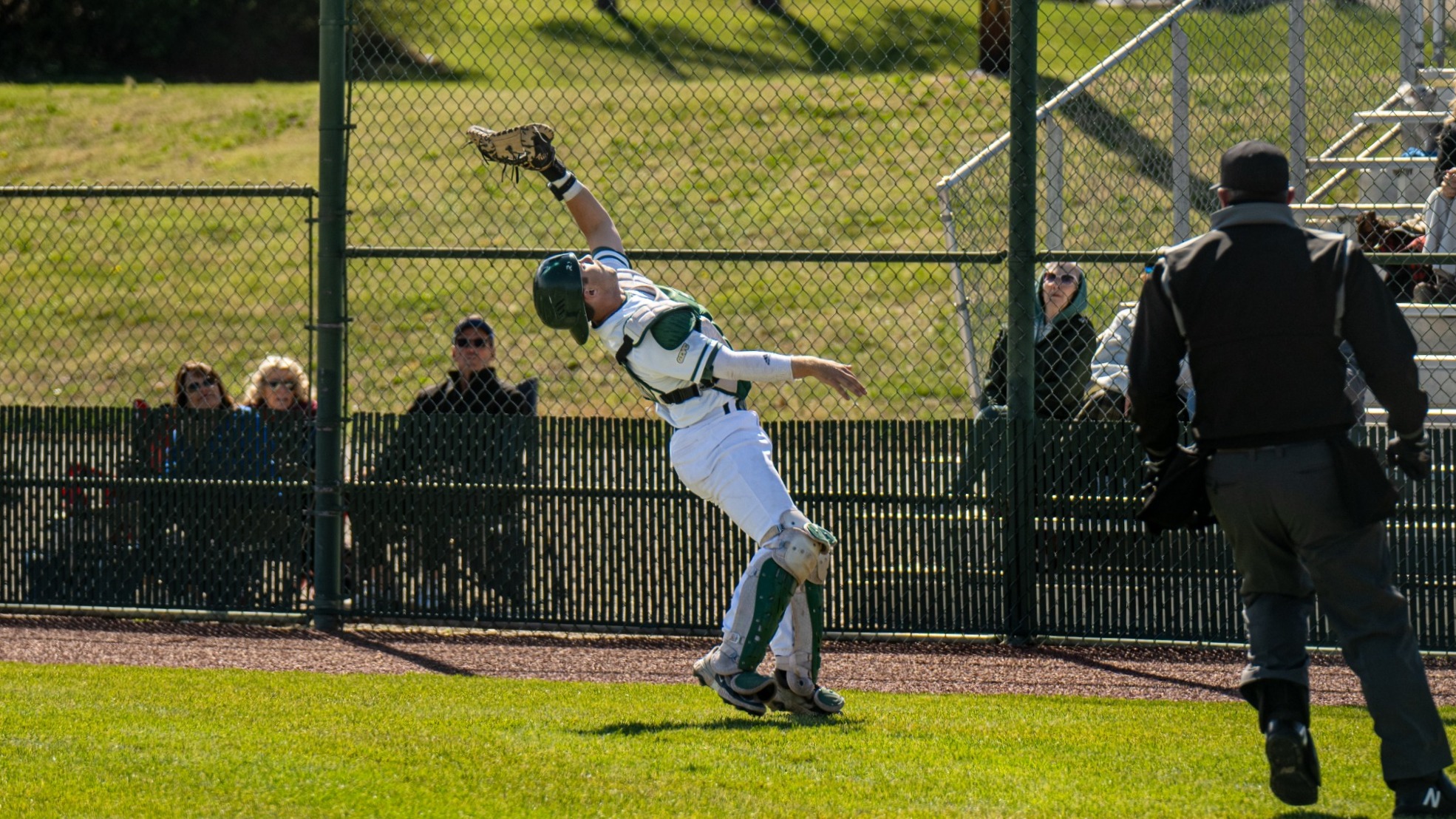 Keaton Scott, wearing full catcher’s gear in a white uniform with green accents, leaps backward near the fence with his glove fully extended overhead while attempting to catch a foul ball along the edge of the field, as an umpire runs in from the right and spectators watch from behind the chain-link fence in the background