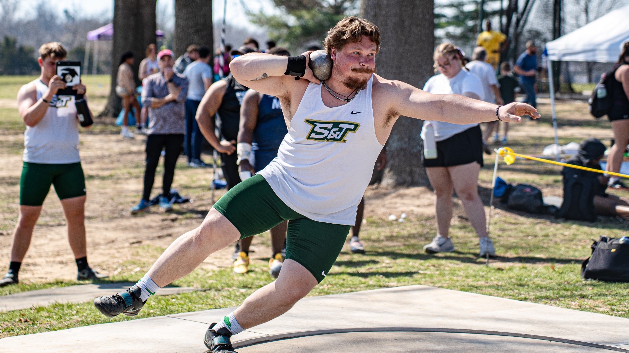 Missouri S&T thrower Tyler Marchert drives forward during a shot put attempt at the Miner Invitational, extending his arm to release the shot while competing athletes and spectators look on.