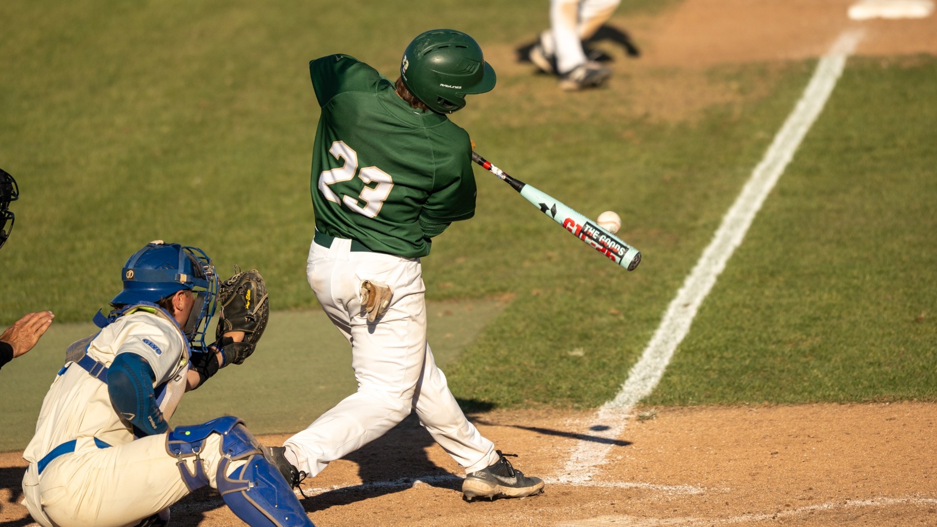 Landen Roberts, wearing jersey number 23 for the Missouri S&T Miners, swings at a pitch during a baseball game against the Rockhurst Hawks. The batter is captured mid-swing with the bat making contact with the ball, while the catcher in blue gear kneels behind him ready to receive the pitch. Another player is visible in the background on the dirt infield.