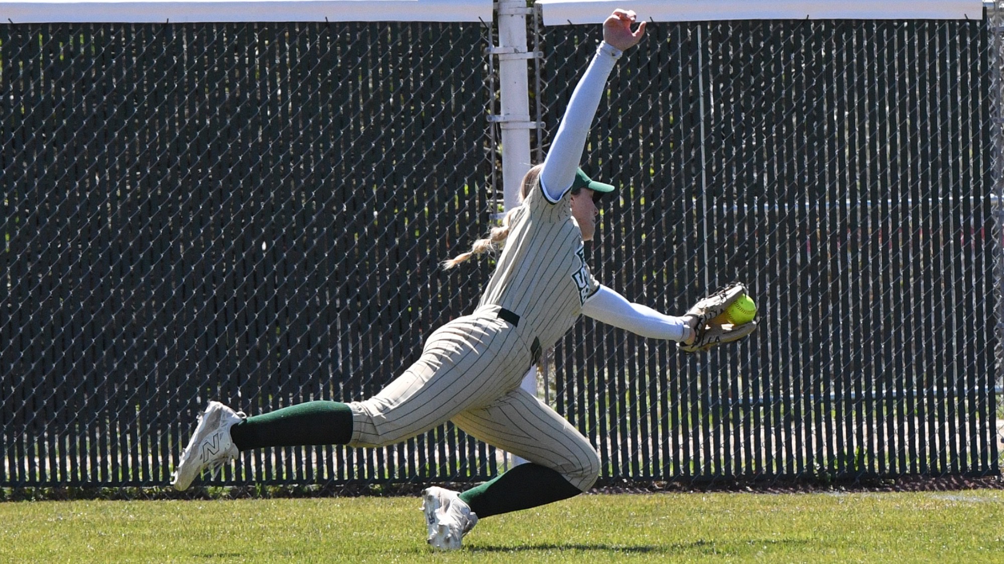 Ty Neal, a Missouri S&T softball right fielder, is photographed sprinting and fully extending her glove hand to make a running catch on a fly ball. She wears a tan pinstriped uniform with green socks and a visor, with her body stretched forward mid-stride as the ball meets her glove near the outfield fence in the background.