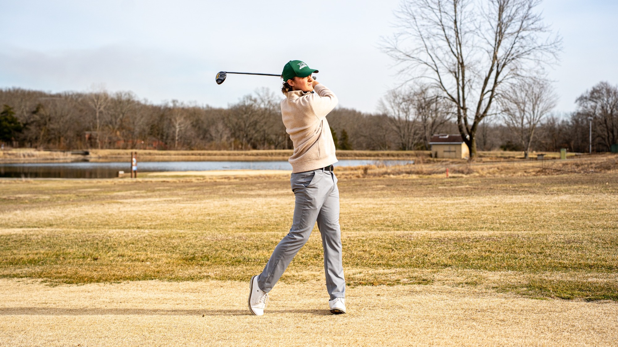 Thomas Heisner, wearing a green cap, beige sweater, and gray pants, follows through on a golf swing on a fairway. He is captured mid-motion with the golf club extended behind his head after striking the ball. The background shows a sunny golf course with dry grass, a pond, bare trees, and a small building in the distance