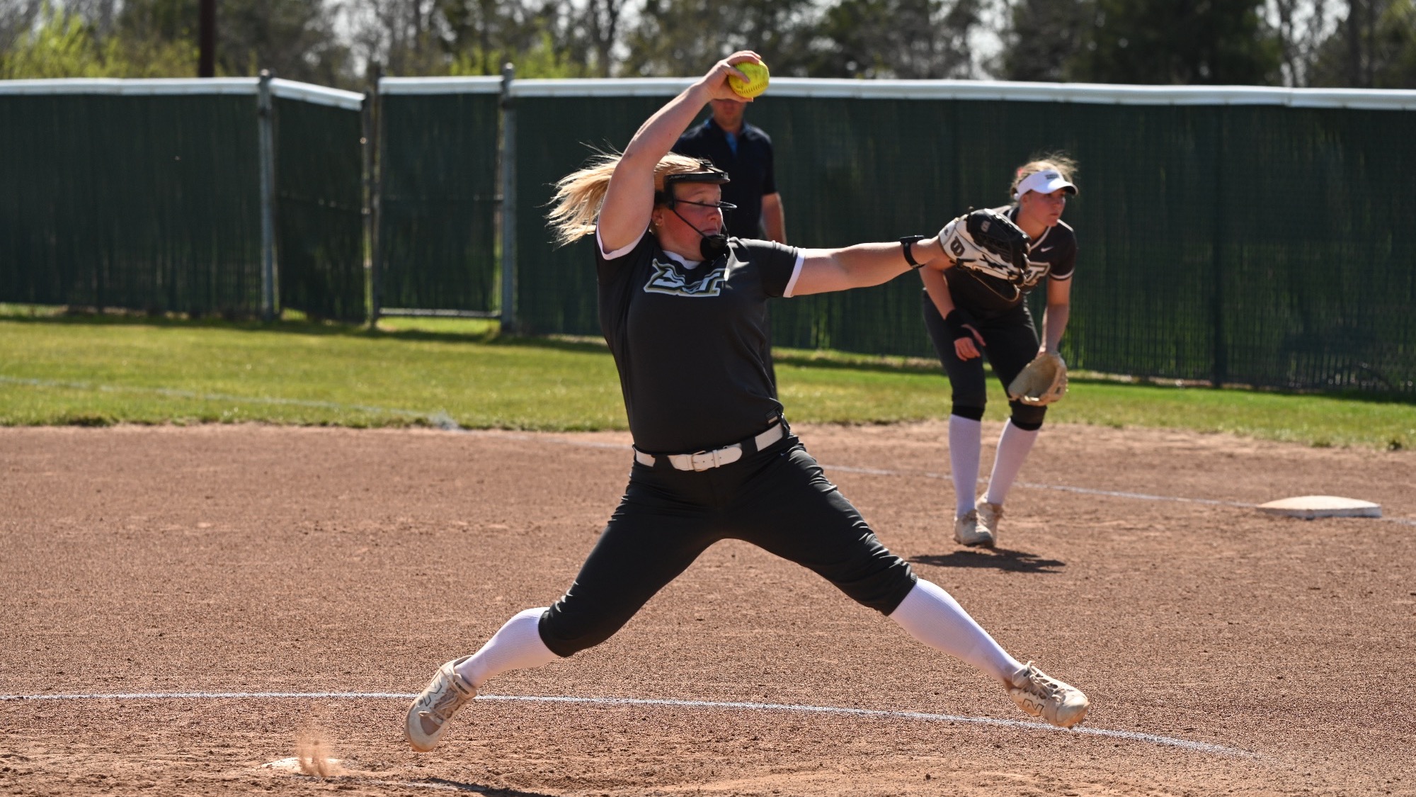 Mia Miller, pitcher for the Missouri S&T Miners, mid-pitch on the dirt mound. She strides forward with legs wide, right arm raised high holding a yellow softball, left arm extended with her glove. She wears a gray Miners jersey, gray pants, white belt and socks, and a face mask. A teammate in gray uniform stands ready in the background on a sunny day.