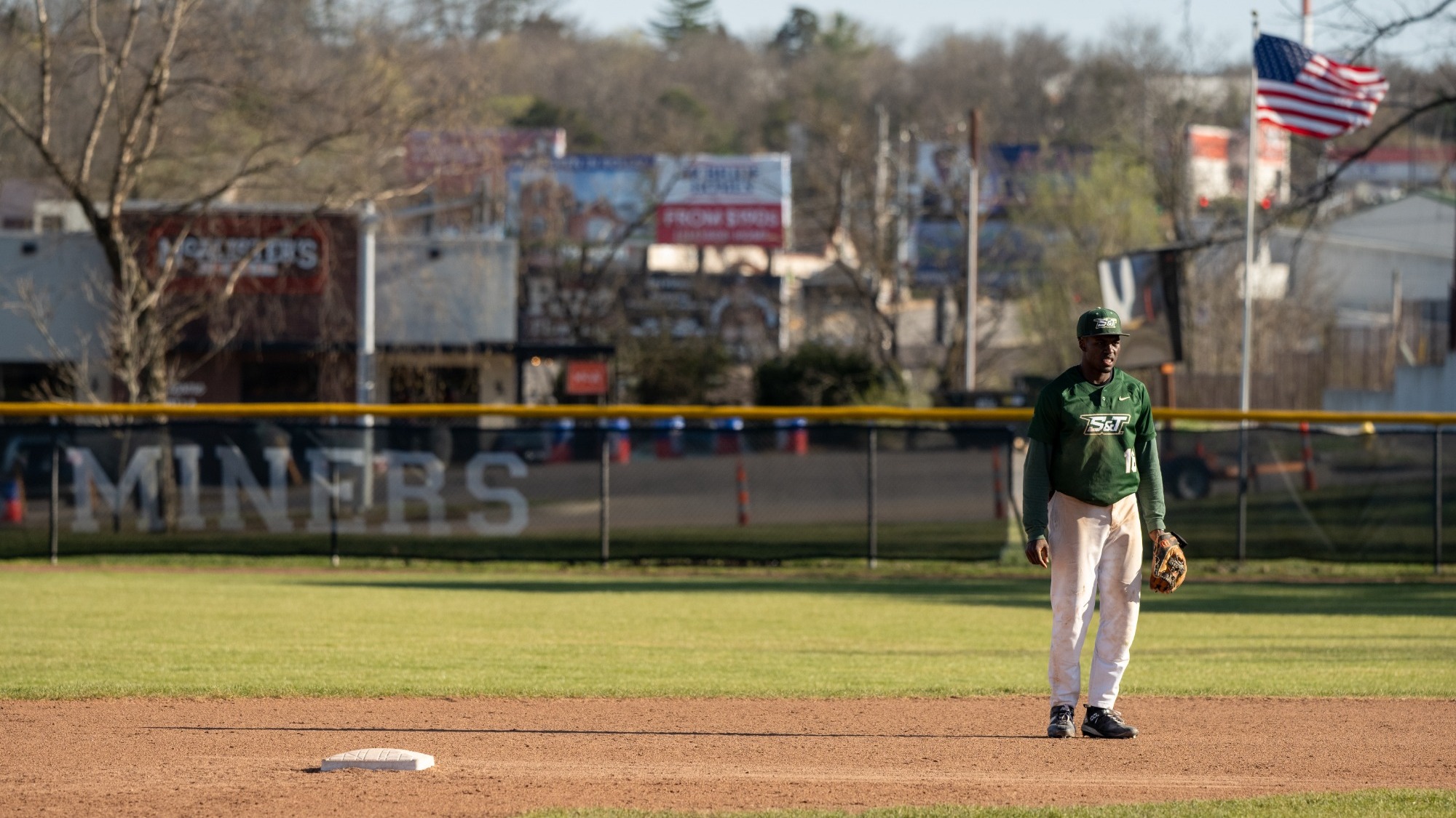 Bricen Smith, a baseball player in a green Missouri S&T Miners jersey number 16, stands on the dirt near first base while holding a glove during a game. An American flag flies in the background.