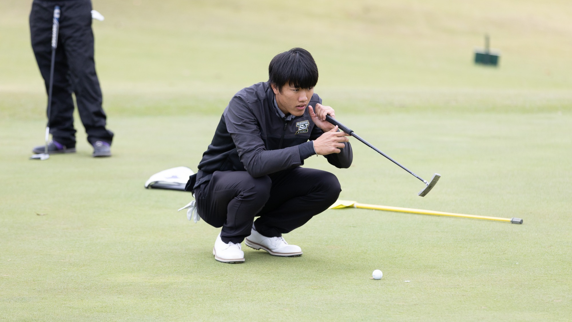 Andy Su is photographed crouching low on the putting green, studying the line of his putt while holding his putter in front of him. He wears a black Missouri S&T jacket, black pants, and white golf shoes, with a golf ball positioned a few feet in front of him on the green. Another golfer stands blurred in the background.