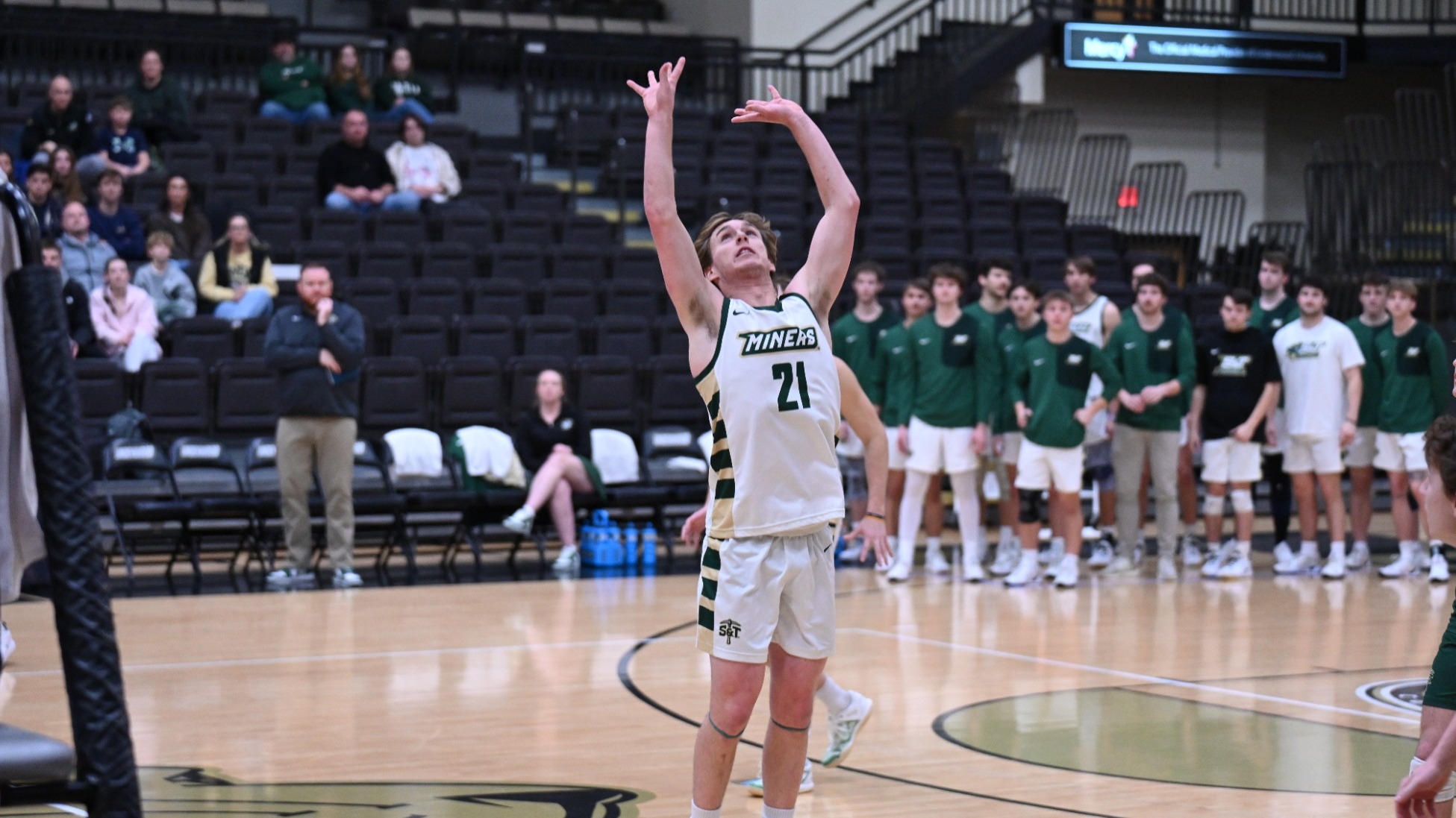 Grant Edmonds is photographed midair in a Missouri S&T basketball uniform, extending both arms upward as he releases a shot near the basket inside Gibson Arena. Teammates and coaches watch from the bench in the background, with the Miners logo visible on the court beneath him.