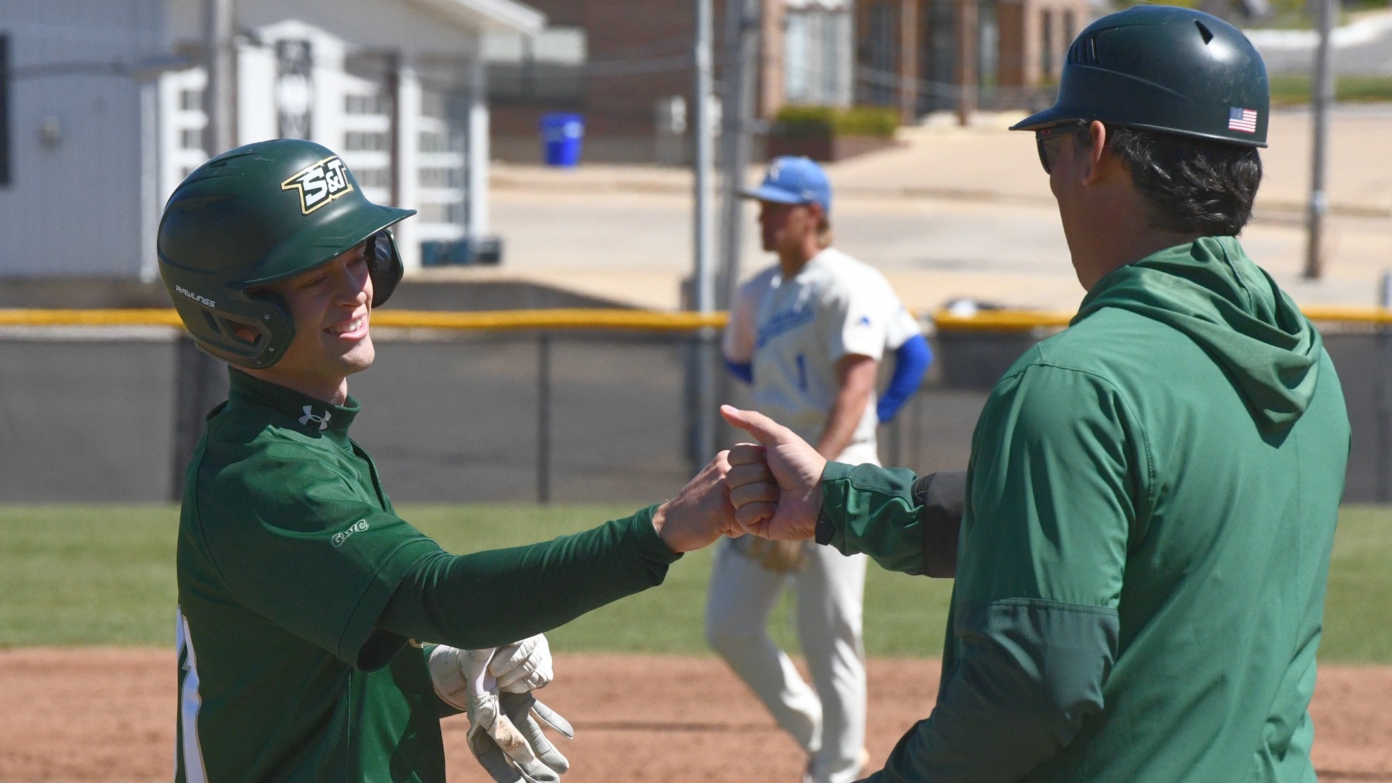 Will Muench, wearing a green Missouri S&T batting helmet and jersey, smiles as he exchanges a fist bump with Assistant Coach Cody Gardner along the first-base line during a game, while an opposing player stands blurred in the background on the infield under sunny conditions
