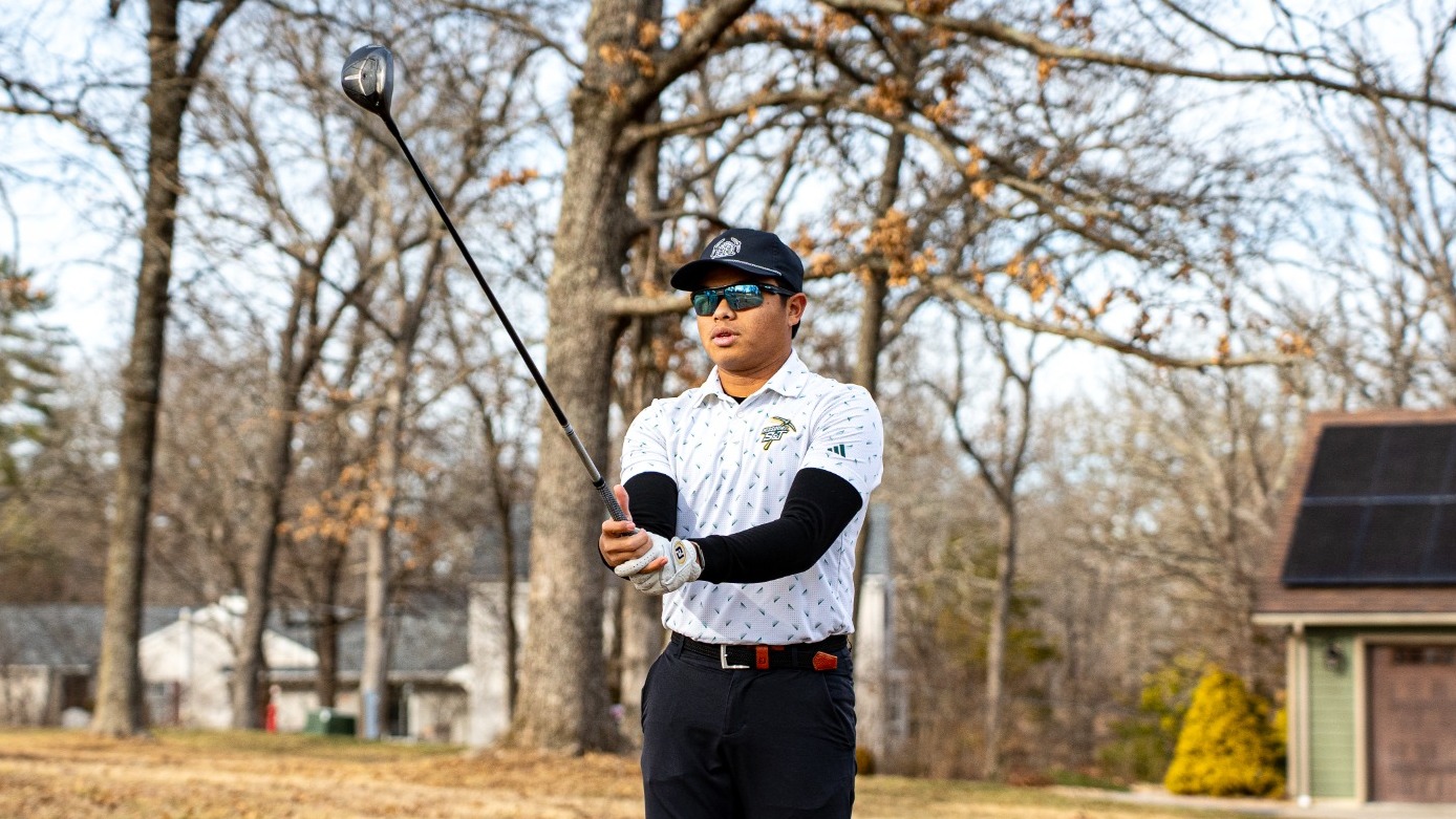 Rizq Adam Rohizam stands on a golf course holding a driver out in front of him after a swing, watching the ball’s flight. He wears a white patterned Missouri S&T polo, black pants, a black cap, sunglasses, and a golf glove, with leafless trees and a small building in the background on a winter-colored fairway