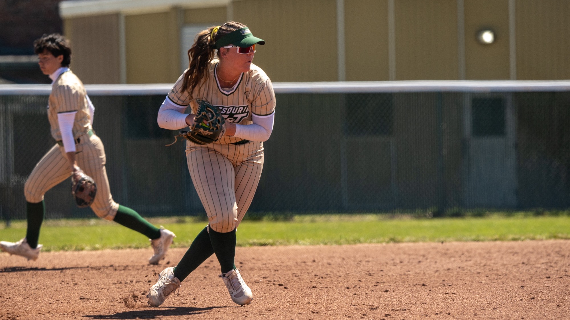 Molly Fitzgerald is photographed in the infield, moving to her left with her glove set by in front of her chest as she fields a ground ball. She wears a tan Missouri S&T uniform with green accents, a green visor, and sunglasses. Another infielder is visible in the background shifting toward the play, with a dirt infield and outfield fence behind them under bright daylight.