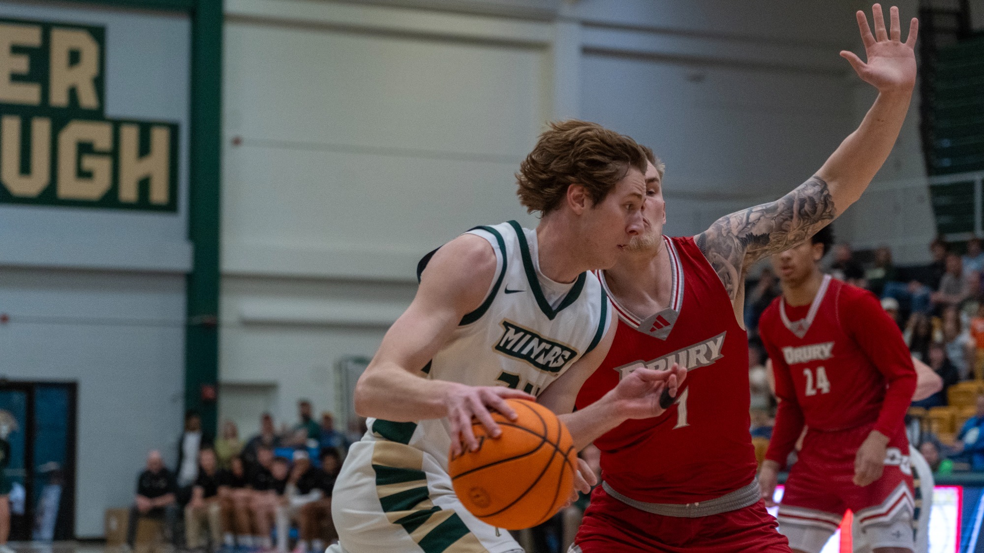 Missouri S&T men’s basketball player Andrew Young is photographed driving toward the basket while dribbling with his right hand, closely guarded by a Drury defender in a red jersey with his arm raised. Another Drury player stands in the background, with fans and gymnasium seating visible behind the play.
