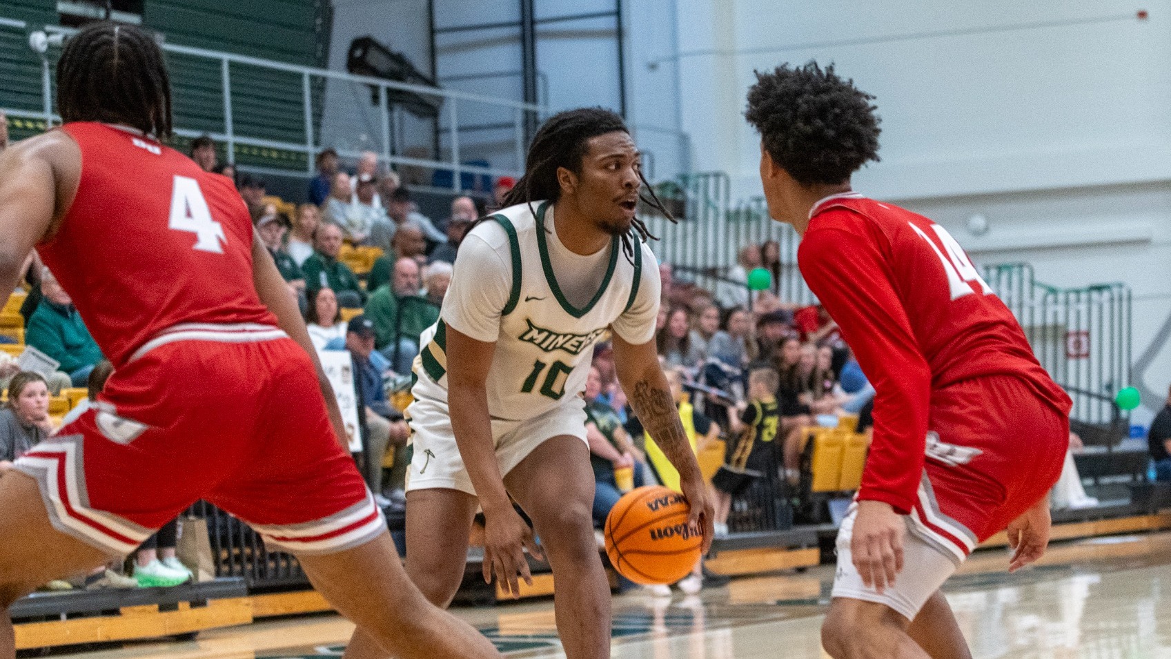 Missouri S&T basketball player Cameron Stovall, wearing a white No. 10 jersey, dribbles the ball near the top of the key while being closely defended by two opponents in red uniforms during a game inside Gibson Arena, with spectators seated in the bleachers in the background.
