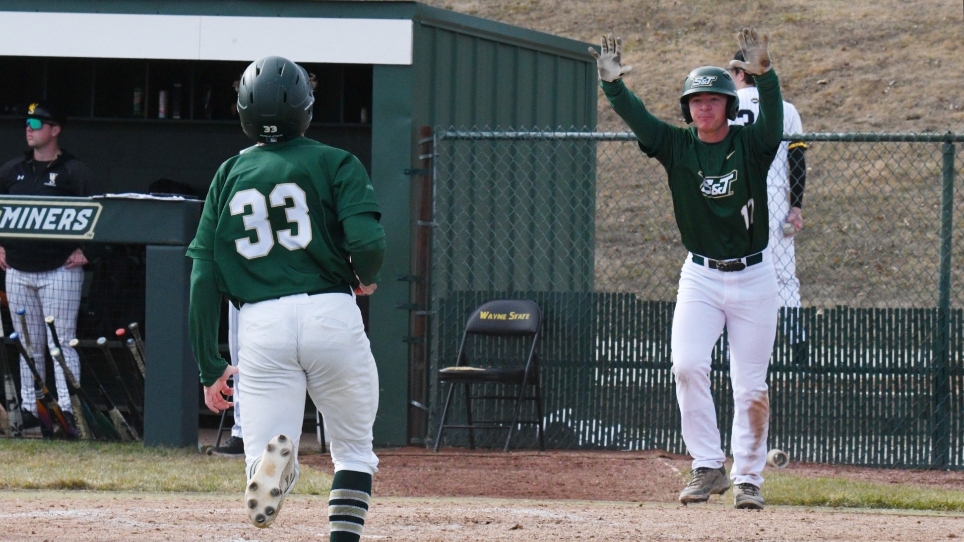 Missouri S&T baseball player Caleb Horsey (#33) runs toward home base while teammate Jackson Evans (#17) raises both hands to signal to Horsey during the play. Evans, wearing a green Miners jersey and batting gloves, is photographed near the dugout entrance preparing to greet Horsey as he approaches, with bats, teammates, and the opposing team dugout visible in the background.