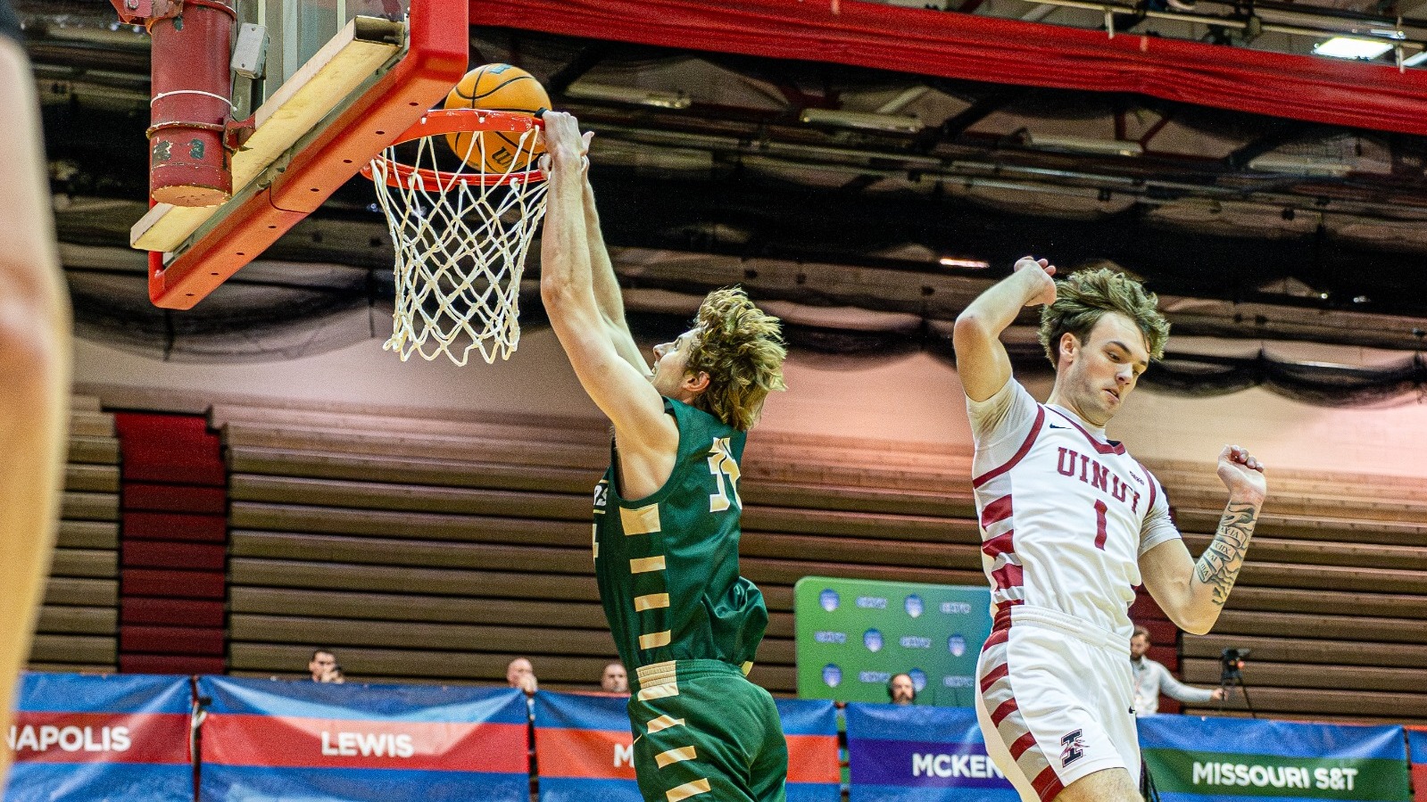Andrew Young of Missouri S&T is photographed leaping toward the rim and finishing a two-handed dunk during a men’s basketball game, with a defender from Indianapolis jumping nearby as the ball goes through the hoop inside a red-seated arena.