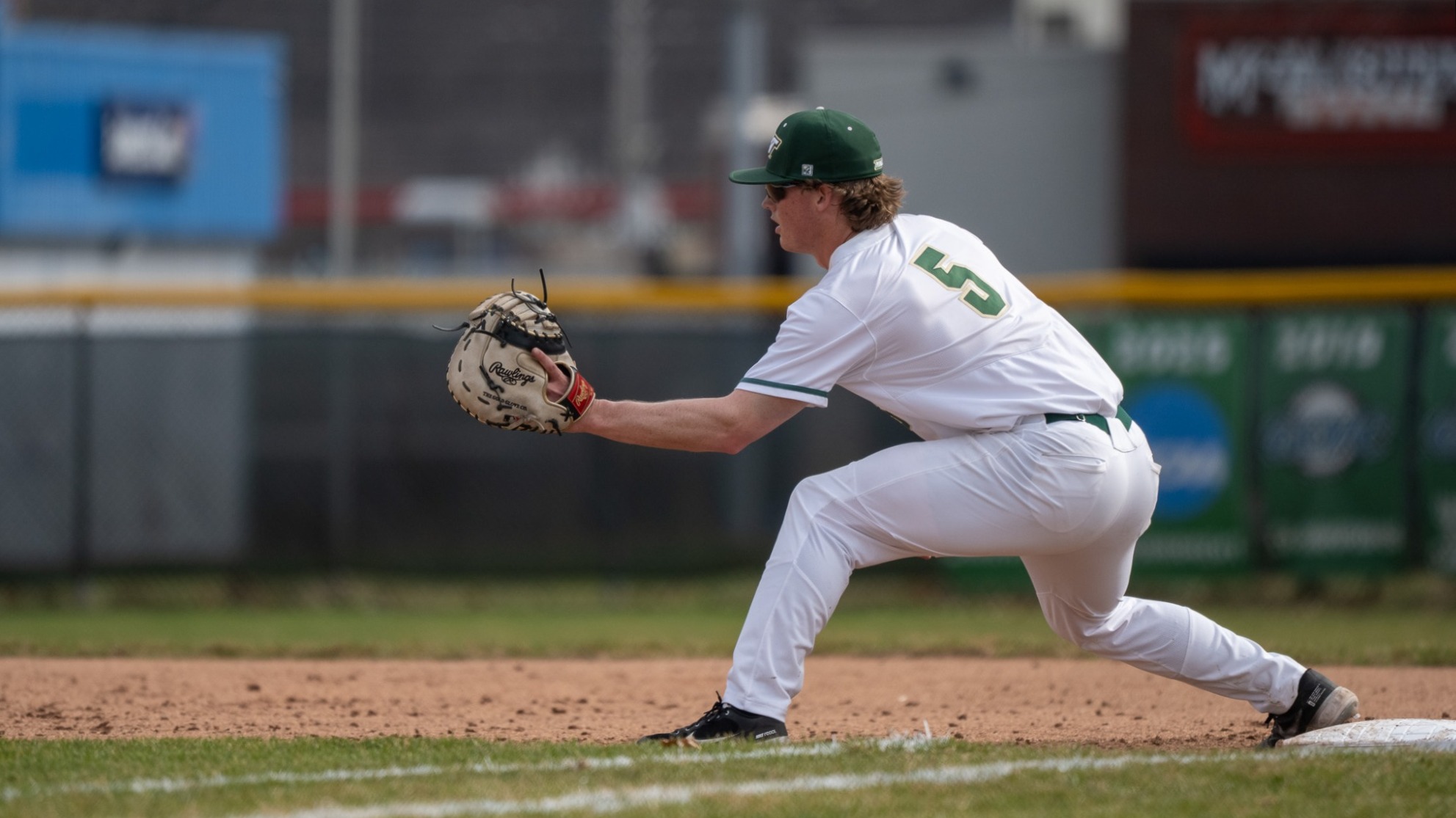 Denton Damgaard is photographed playing first base for Missouri S&T, positioned in a low fielding stance with his glove extended out in front while preparing to receive a throw at first. He is wearing a white Miners uniform and green cap, with one foot on the first base bag and the infield dirt and outfield fence visible in the background.