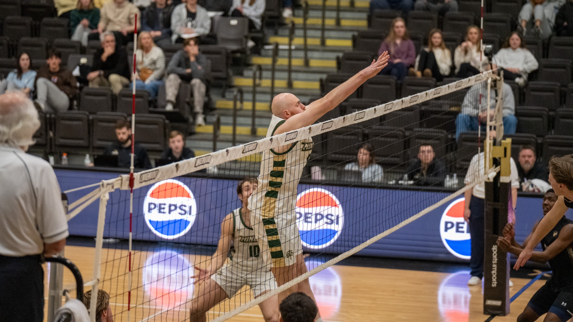 Caleb Rohlwing, a Missouri S&T volleyball player in a white and green jersey, jumps high at the net to block a shot during a collegiate match in a crowded arena.