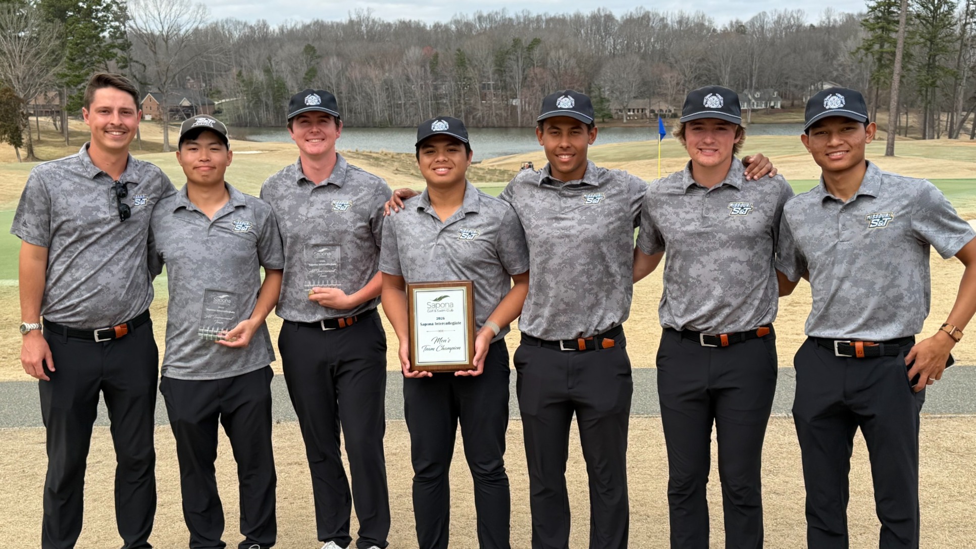 A group portrait of the Missouri S&T men’s golf team standing on a grass field at a golf course after winning the Sapona Intercollegiate championship. Seven men—a coach and six players—are dressed in matching grey digital camouflage polo shirts with 
