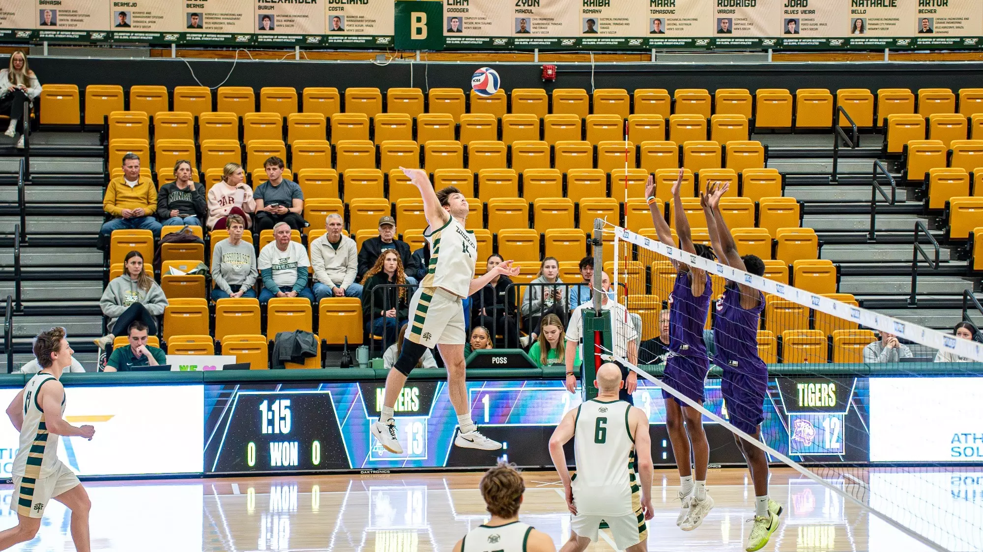 Missouri S&T opposite-side hitter Aaron Sallade in a white Miners jersey elevates at the net for a spike attempt in Gibson Arena while two Edward Waters blockers in purple jump to contest during the match.