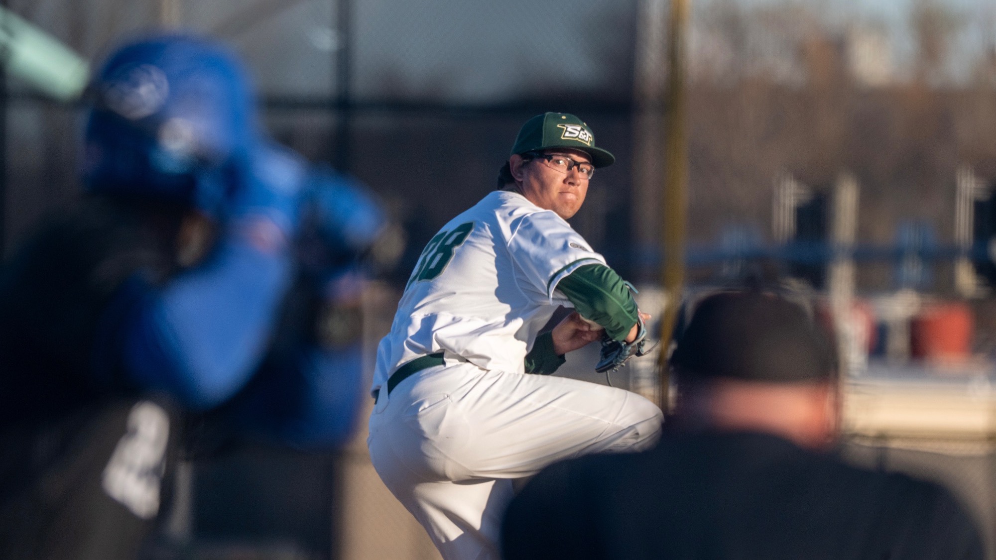 Brandon Hull, a Missouri S&T baseball pitcher, is photographed mid-delivery on the mound, lifting his front leg and preparing to throw a pitch while an umpire and opposing batter appear blurred in the foreground.