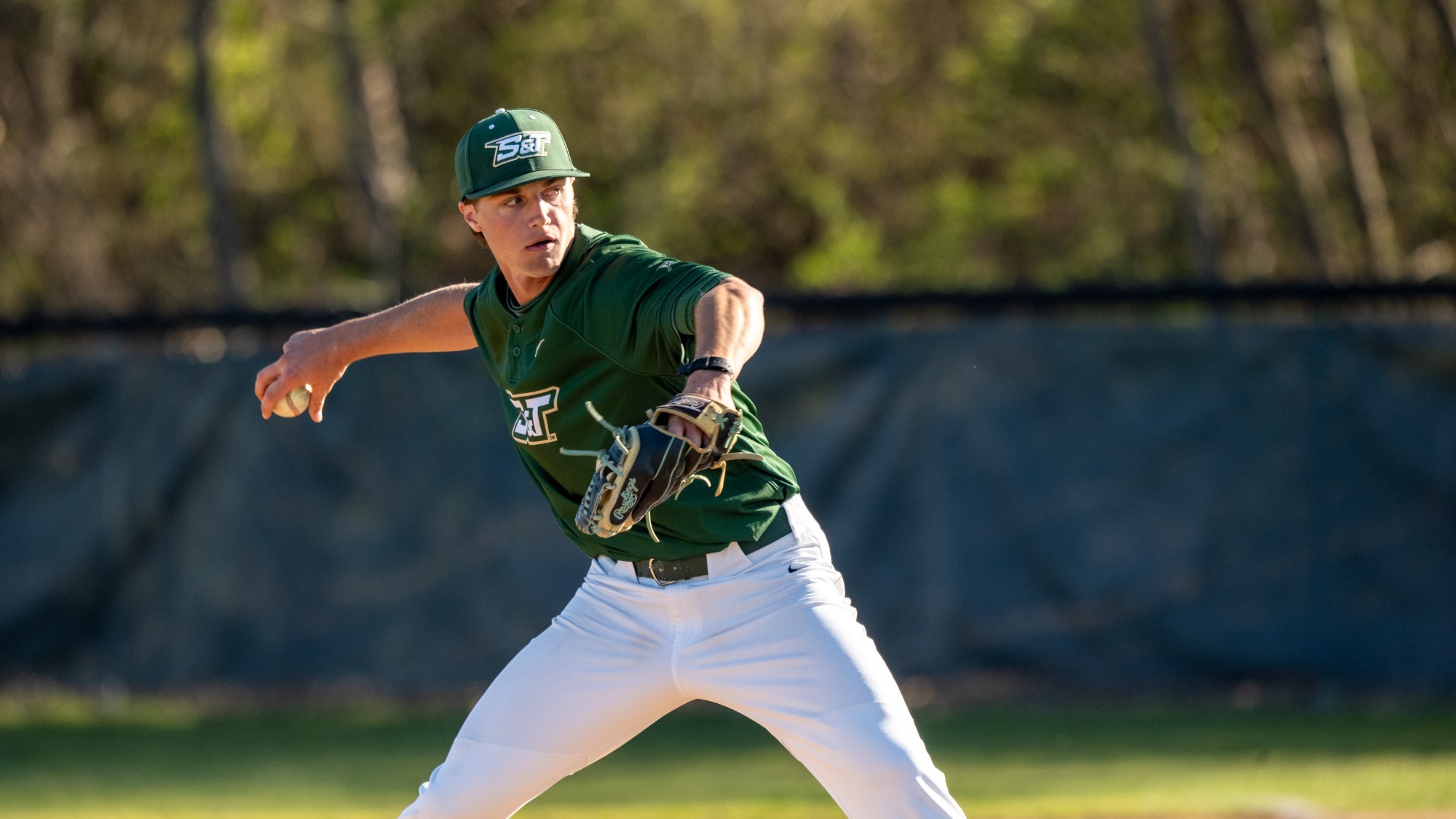 Missouri S&T baseball player Nate Donovan, wearing a green Miners jersey and cap with white pants, delivers a pitch from the mound during a game. He is mid-throw with his right arm extended back holding the baseball and his glove hand forward, focused toward home plate. The background shows an outfield fence and trees in soft focus under daylight conditions