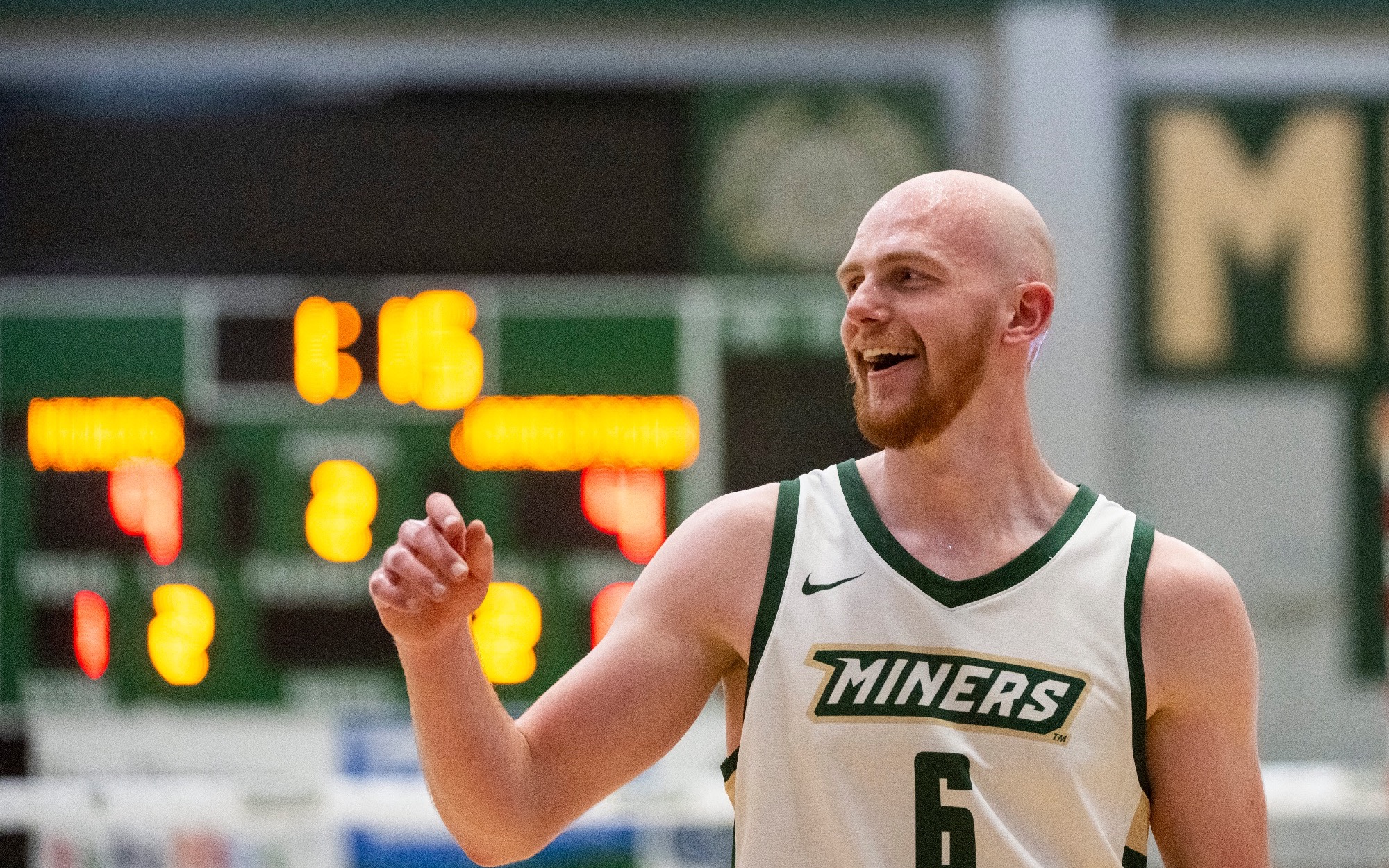 Caleb Rohlwing is photographed on an indoor volleyball court wearing a white Missouri S&T “Miners” jersey with the number 6. He is smiling and gesturing with his right hand, looking off to the side, with a blurred scoreboard and gym background behind him.