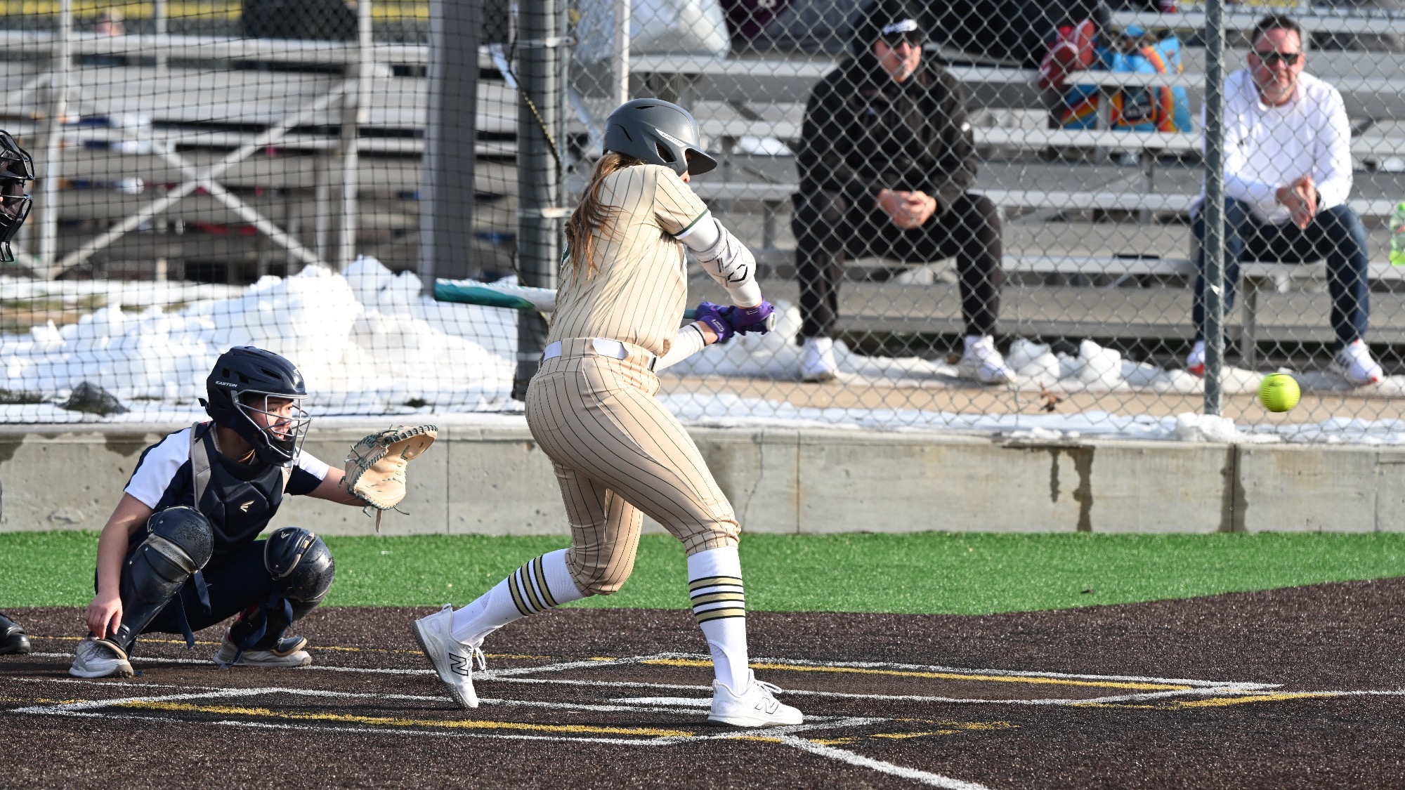 Missouri S&T softball player Morgan Williams, wearing a gold pinstripe Miners jersey, makes contact with the ball at the plate while the catcher sets up behind her during a game.
