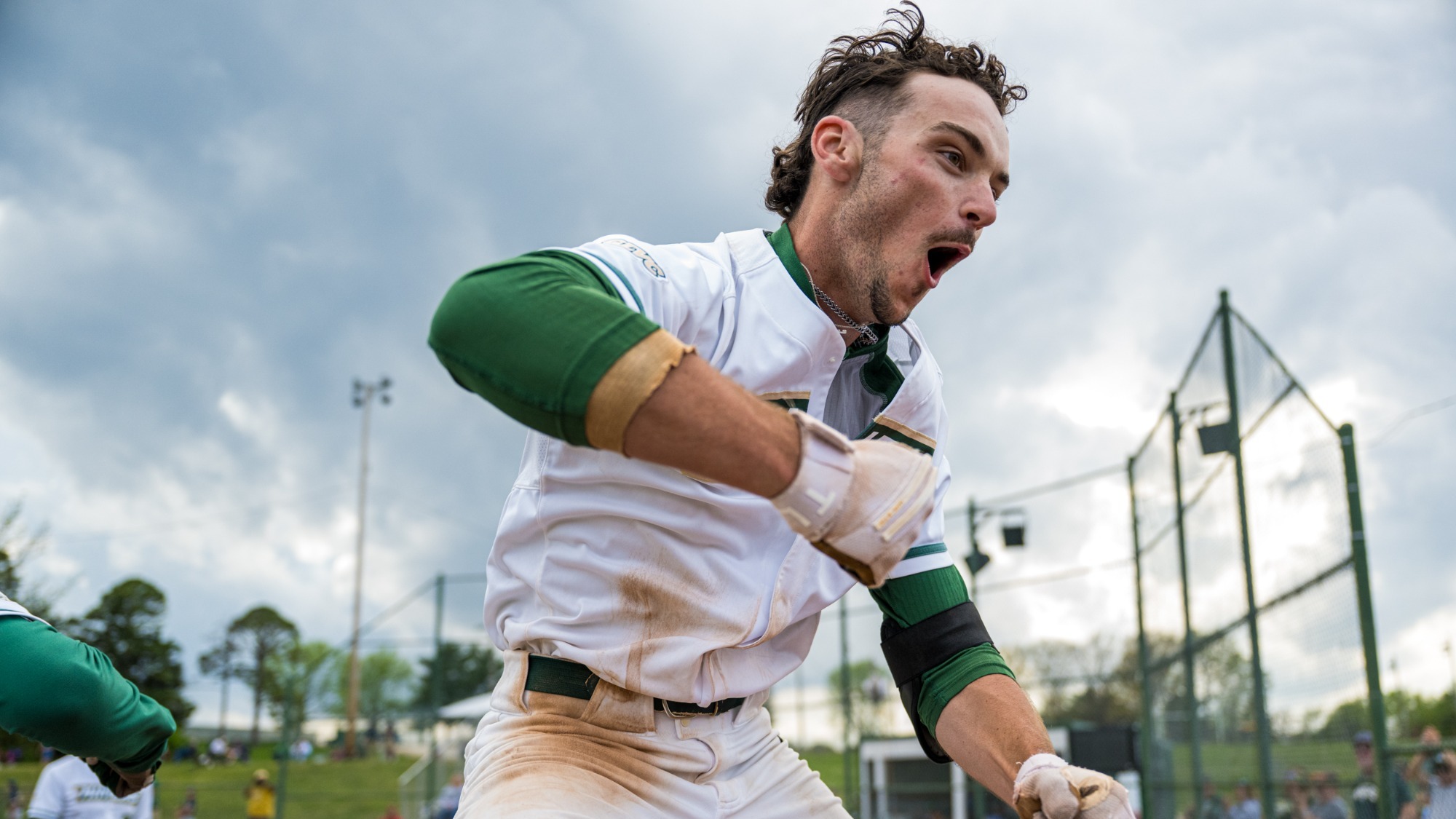 Garrett Meyer of the Missouri S&T Miners celebrates after hitting a home run. The baseball player, wearing a dirt-stained white and green Missouri S&T Miners uniform, has his mouth wide open in an excited shout, both fists clenched and pumped forward, standing on the baseball field with a chain-link fence and spectators visible in the background
