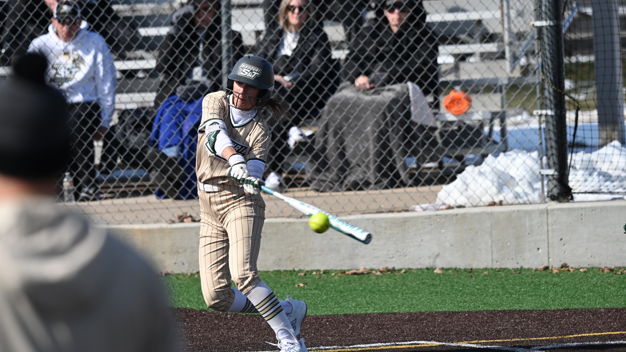 Missouri S&T softball player Kaylee LaChappel, wearing a gold pinstripe Miners jersey, makes contact with the ball during an at-bat as spectators watch from the stands behind the fence.