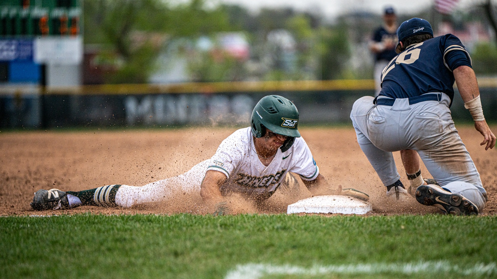 Drew Baugus of Missouri S&T slides headfirst into a base during a game against Illinois Springfield, kicking up a cloud of infield dirt as a defender applies a tag attempt near the bag. Baugus wears a white Miners uniform and green helmet, fully extended as he reaches the base.