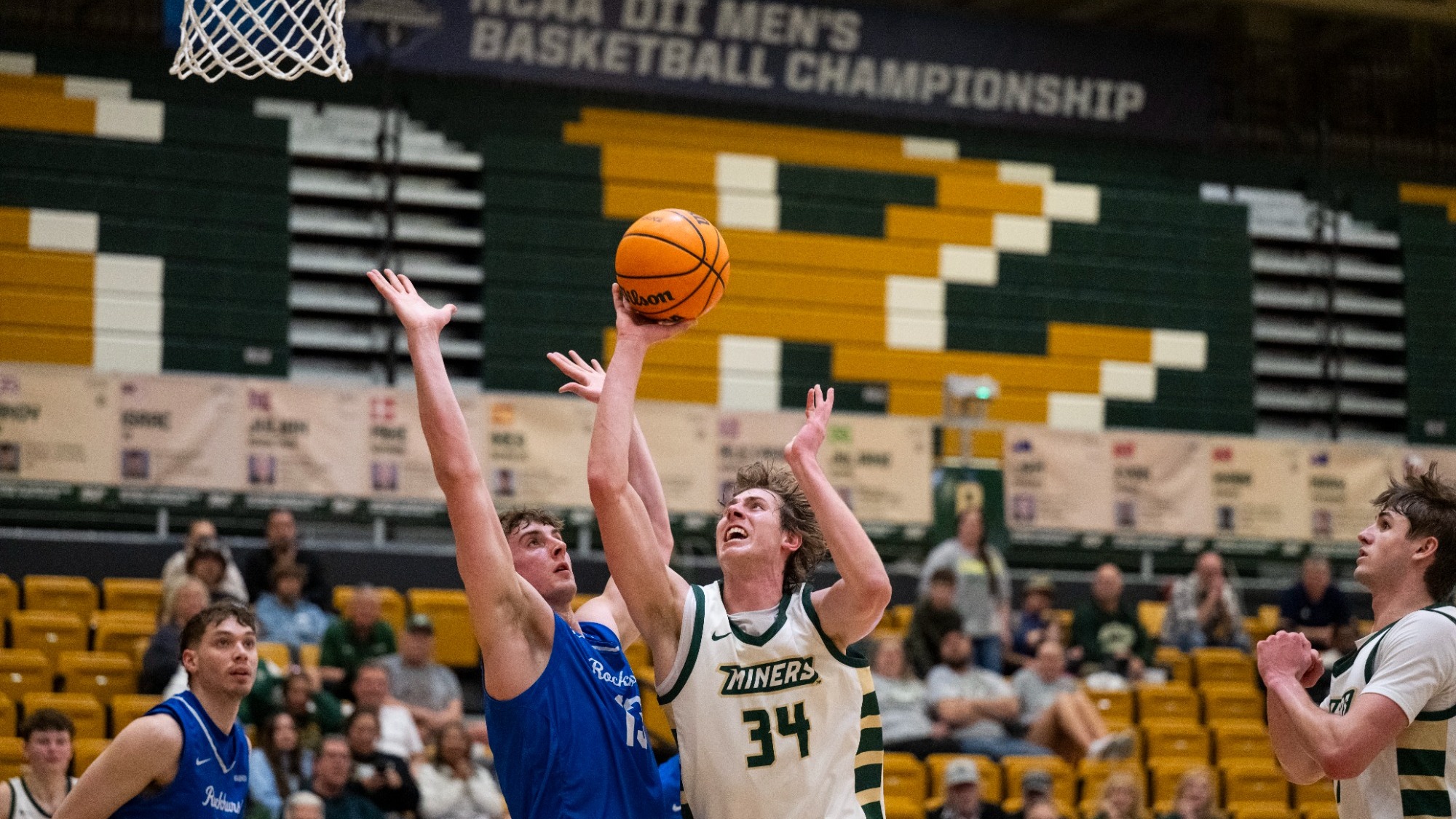 Andrew Young (#34) of the Miners men’s basketball team drives toward the basket for a right-handed layup while closely contested by a Rockhurst defender in a blue jersey with both arms raised. Young is wearing a white Missouri S&T uniform with green and gold trim, “Miners” across the chest, and green side stripes on the shorts. The play happens directly beneath the hoop inside a gym with gold and green seating, with an “NCAA DII Men’s Basketball Championship” banner visible above the court and spectators seated in the background