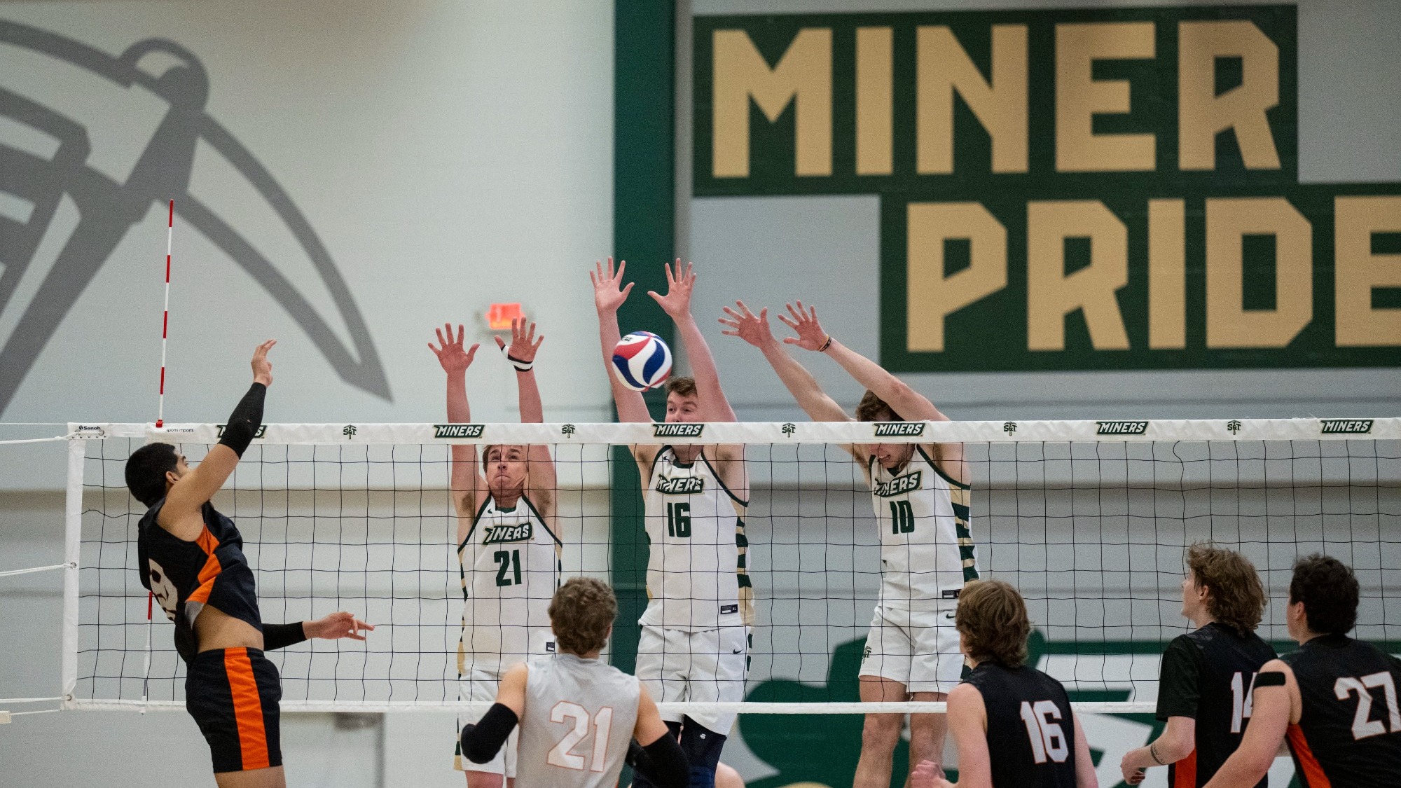 Men’s volleyball action shot at a Miners home match. On the left side of the net, A Jamestown player (wearing black and orange) jumps high with his right arm extended, spiking the volleyball. In the center and right, three Miners players in white jerseys leap to block the spike: Grogan Kraus (#16) in the middle, Nate Meyer (#10) on the right, and Grant Edmonds (#21) on the left.