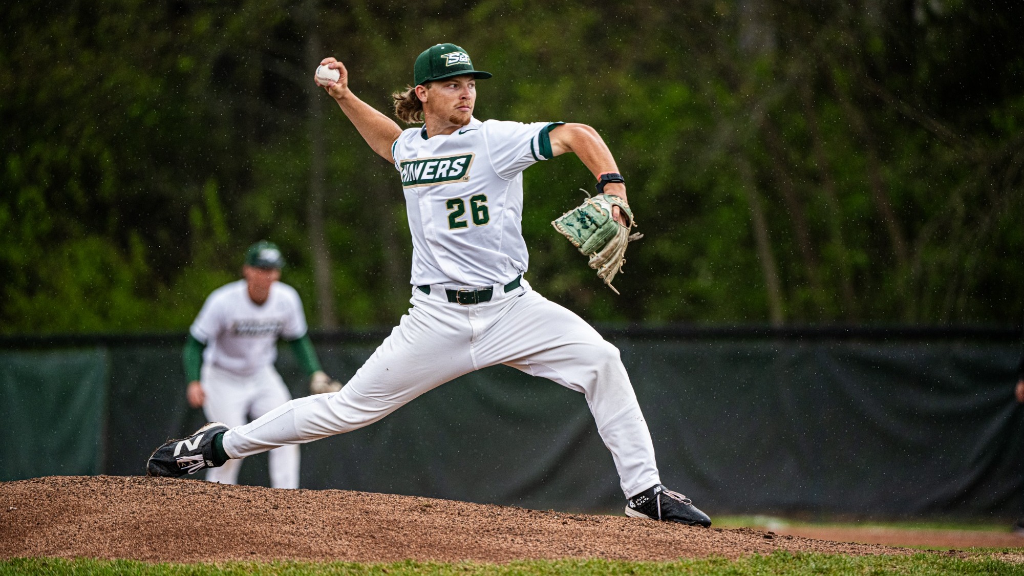 Jake Schisler, a right-handed pitcher for Missouri S&T, delivers a pitch from the mound during light rain while wearing a white Miners uniform with green lettering and the number 26, a green S&T cap, white pants, and black cleats. His stride leg is extended forward and his throwing arm is drawn back mid-motion, with a teammate in a matching white uniform blurred in the background near the infield and trees beyond the outfield fence.