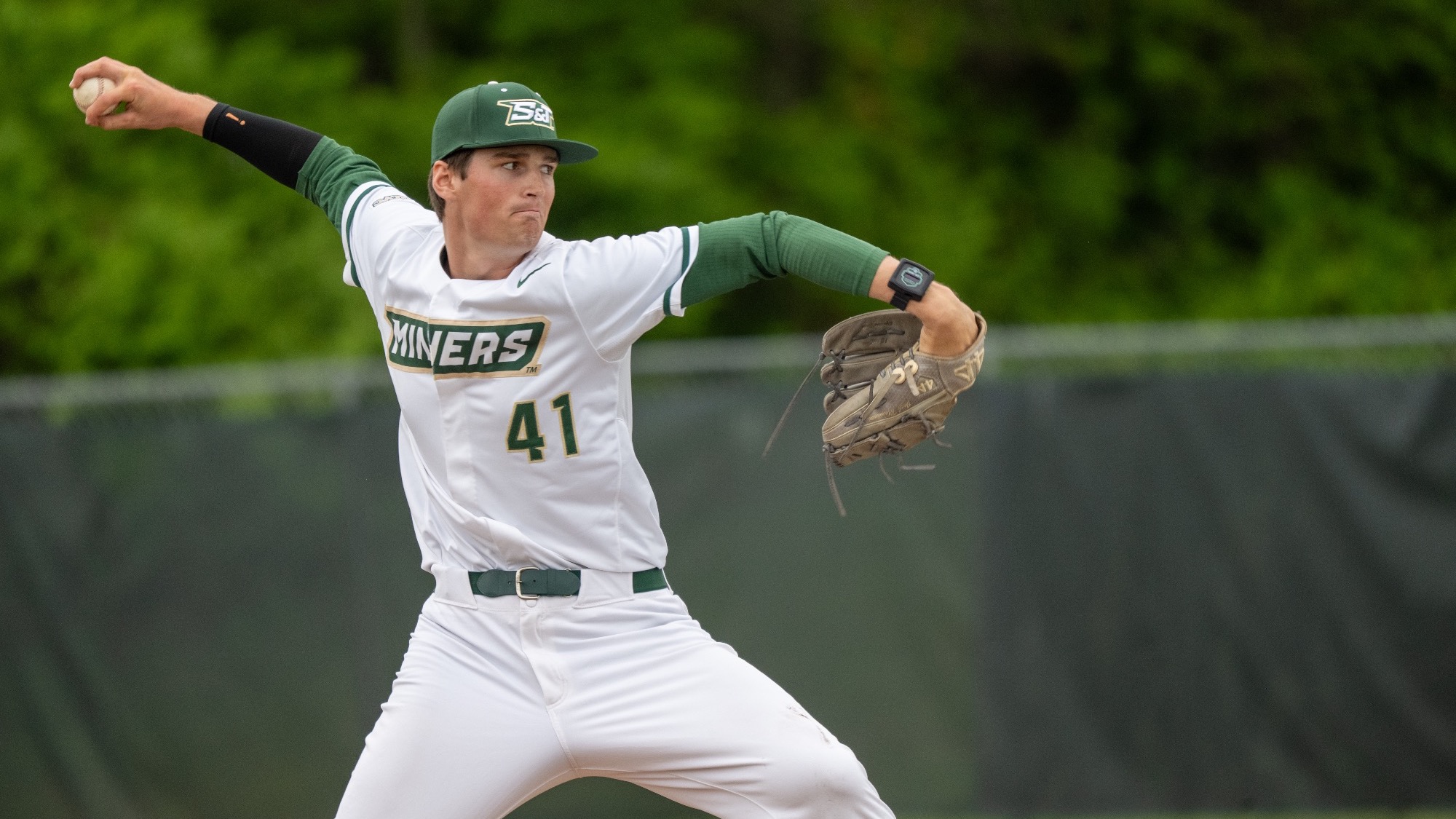 Jack Manninen is photographed on the mound mid-delivery, wearing a white Missouri S&T Miners uniform with green sleeves and cap, his right arm extended back holding the baseball and his left arm forward with the glove, preparing to throw a pitch with a focused expression against a blurred green outfield background.
