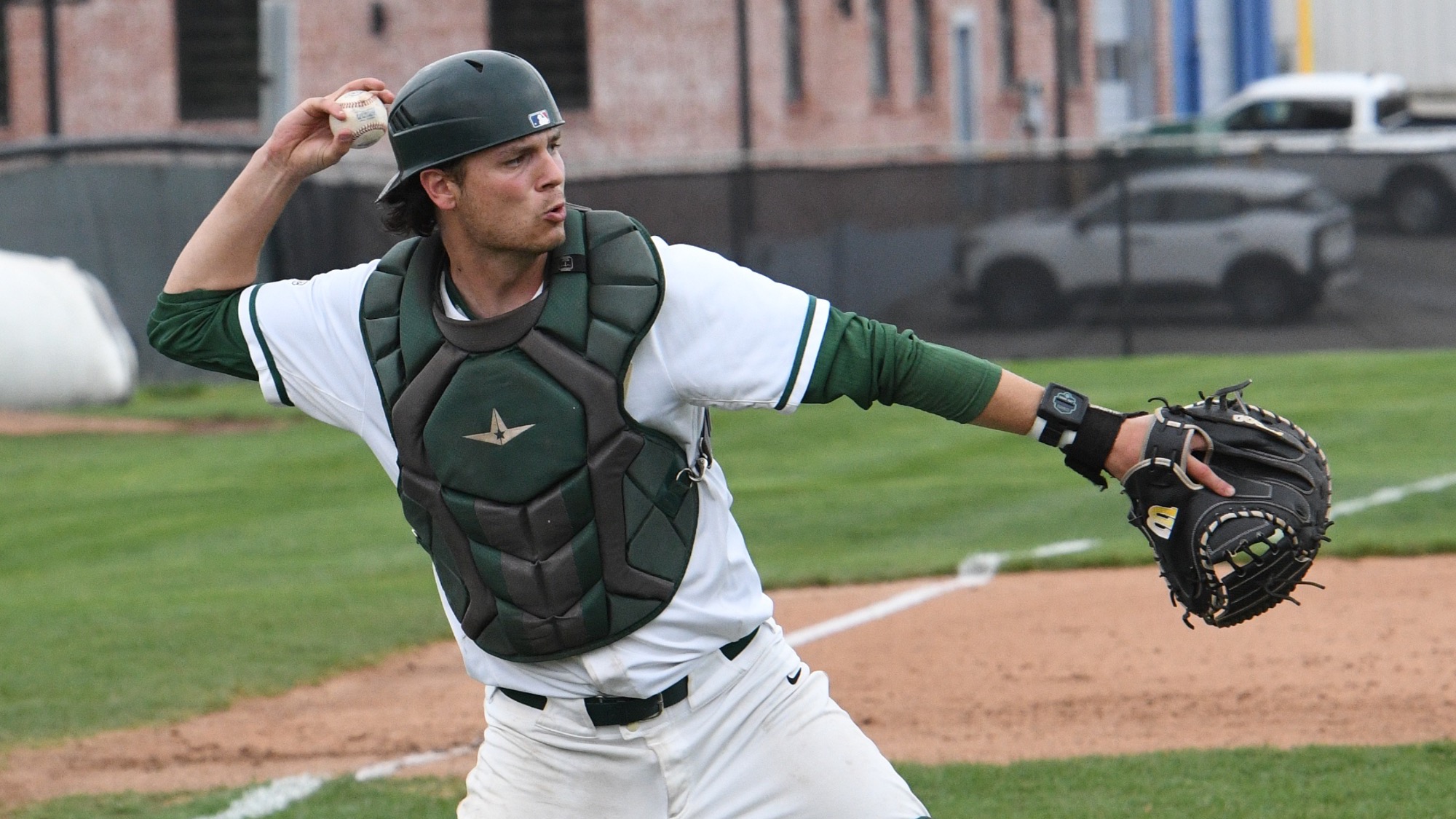 Tyler Bizzle, a baseball catcher, is photographed mid-throw on the field, wearing a white uniform with green sleeves, a dark green chest protector, and a helmet. He has his right arm extended back holding the baseball, preparing to throw, while his left arm reaches forward with a catcher’s mitt
