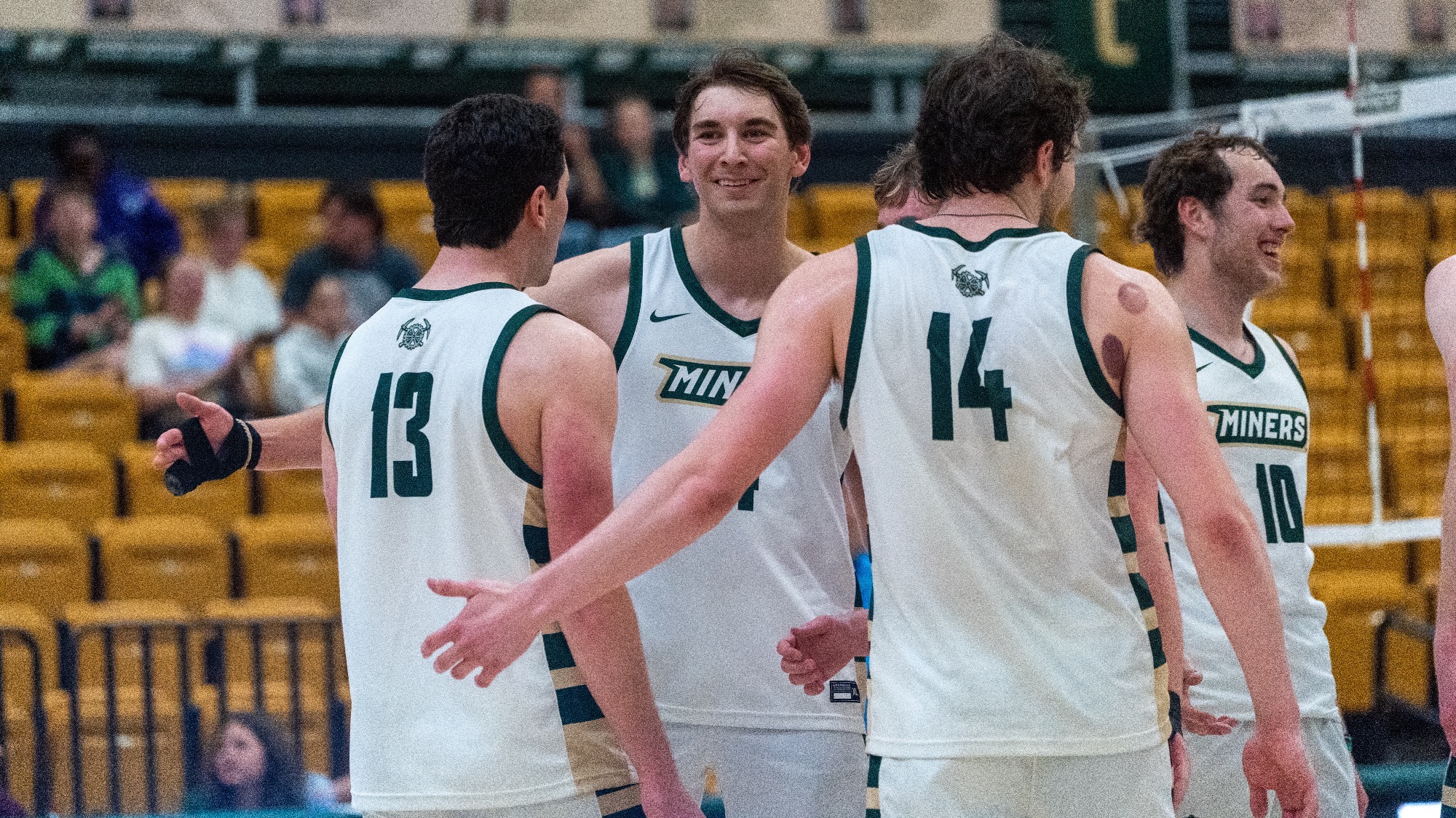Tyler Johnwick is photographed at center, smiling and facing teammates during a break in play on an indoor volleyball court, wearing a white Missouri S&T jersey. Around him, Zachary Carff (#13), Aaron Sallade (#14), and Nate Meyer (#10) are gathered in a small huddle, also in white jerseys with green trim, appearing to celebrate