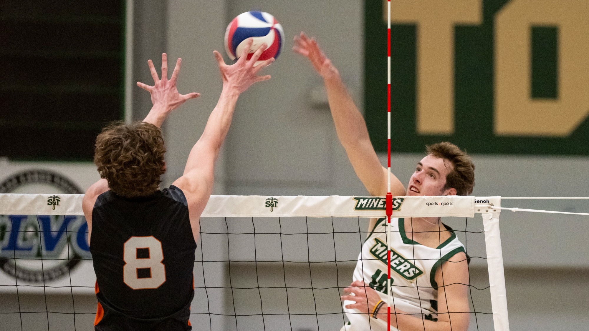 Missouri S&T volleyball player Nate Meyer, wearing a white “Miners” jersey, jumps at the net with his right arm extended to tip the ball over an opposing blocker in a black jersey (#8).