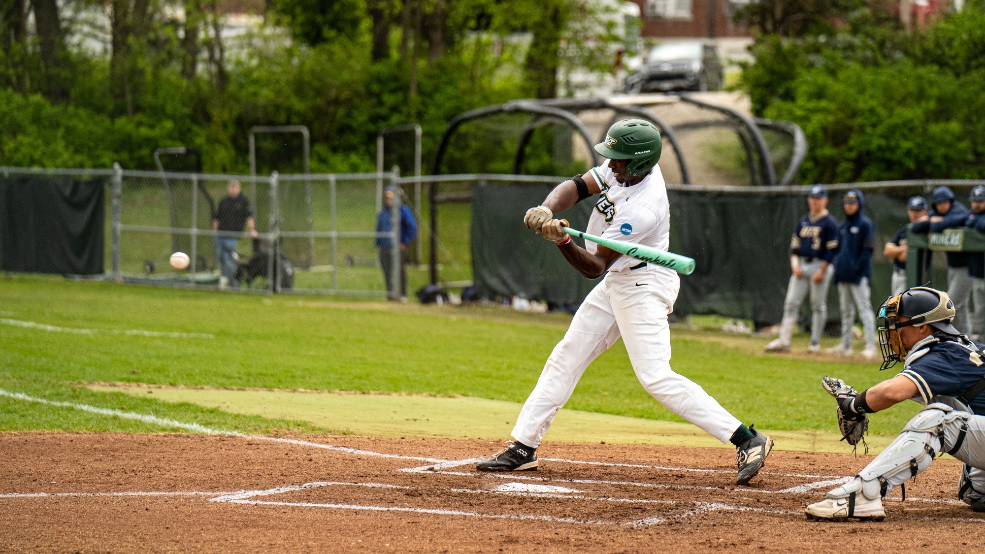 Bricen Smith of Missouri S&T swings through a pitch in a white jersey at the Missouri S&T baseball field, making contact at home plate as the catcher sets behind him.