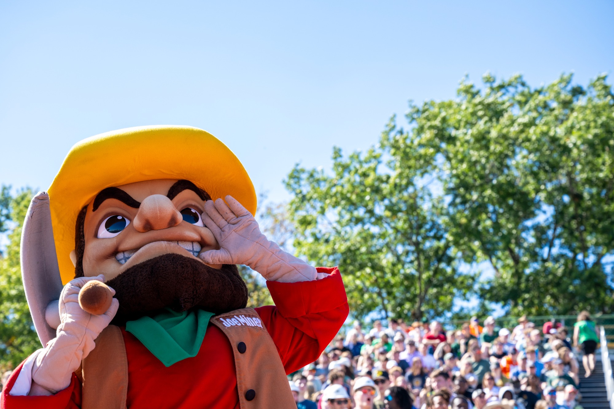 A photo looking up at Joe Miner as the mascot stands with his hand to his mouth and a pickaxe over his shoulder. Clear skies, a tree and crowded stands full of fans are visible in the background. Joe wears a red shirt, a brown vest, a green bandana, and a yellow hat