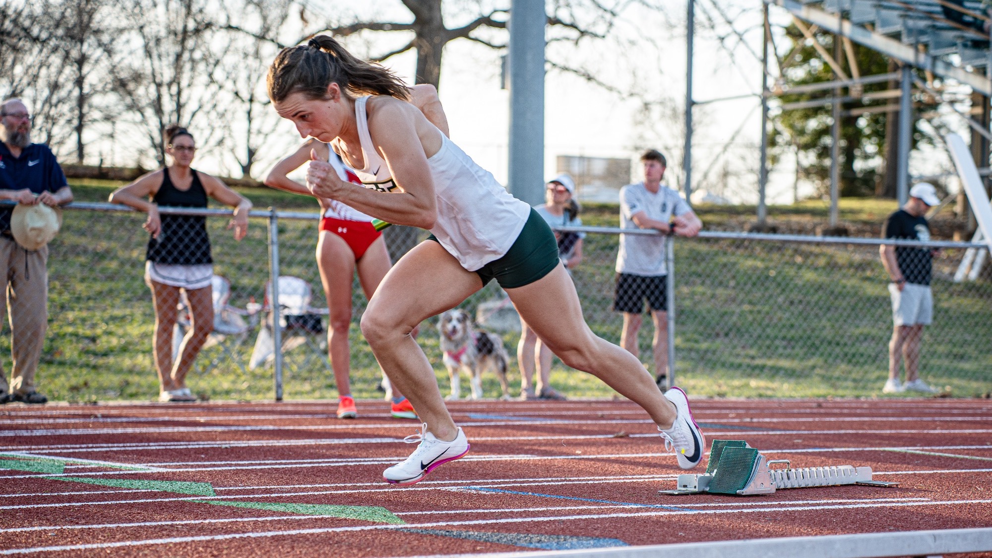 Missouri S&T runner Mary Kimmerle explodes out of the starting blocks to begin her leg of the 4x400-meter relay at the Miner Invitational, driving forward on the track as spectators watch from behind the fence.