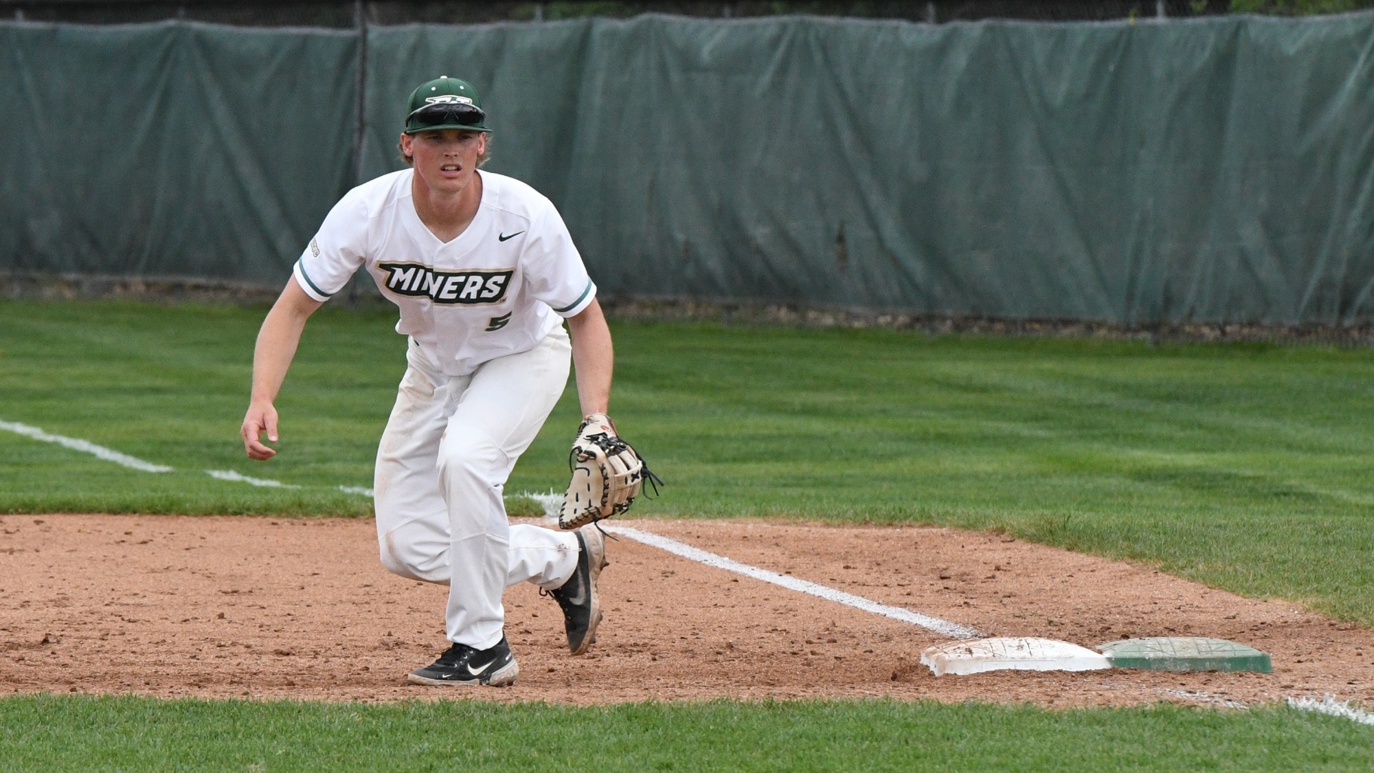 Denton Damgaard photographed wearing a white S&T jersey with green trim and a green baseball cap. He is standing near first base and covering it during an opposing team's at bat.