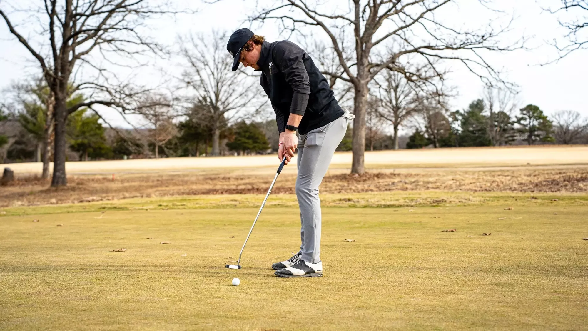 Chayton Jordan of Missouri S&T Men's Golf is photographed mid-putt, with his club in motion and on his follow through after striking the ball. He is wearing a black quarter zip and gray pants with Missouri S&T logos on his baseball cap and on his quarter zip