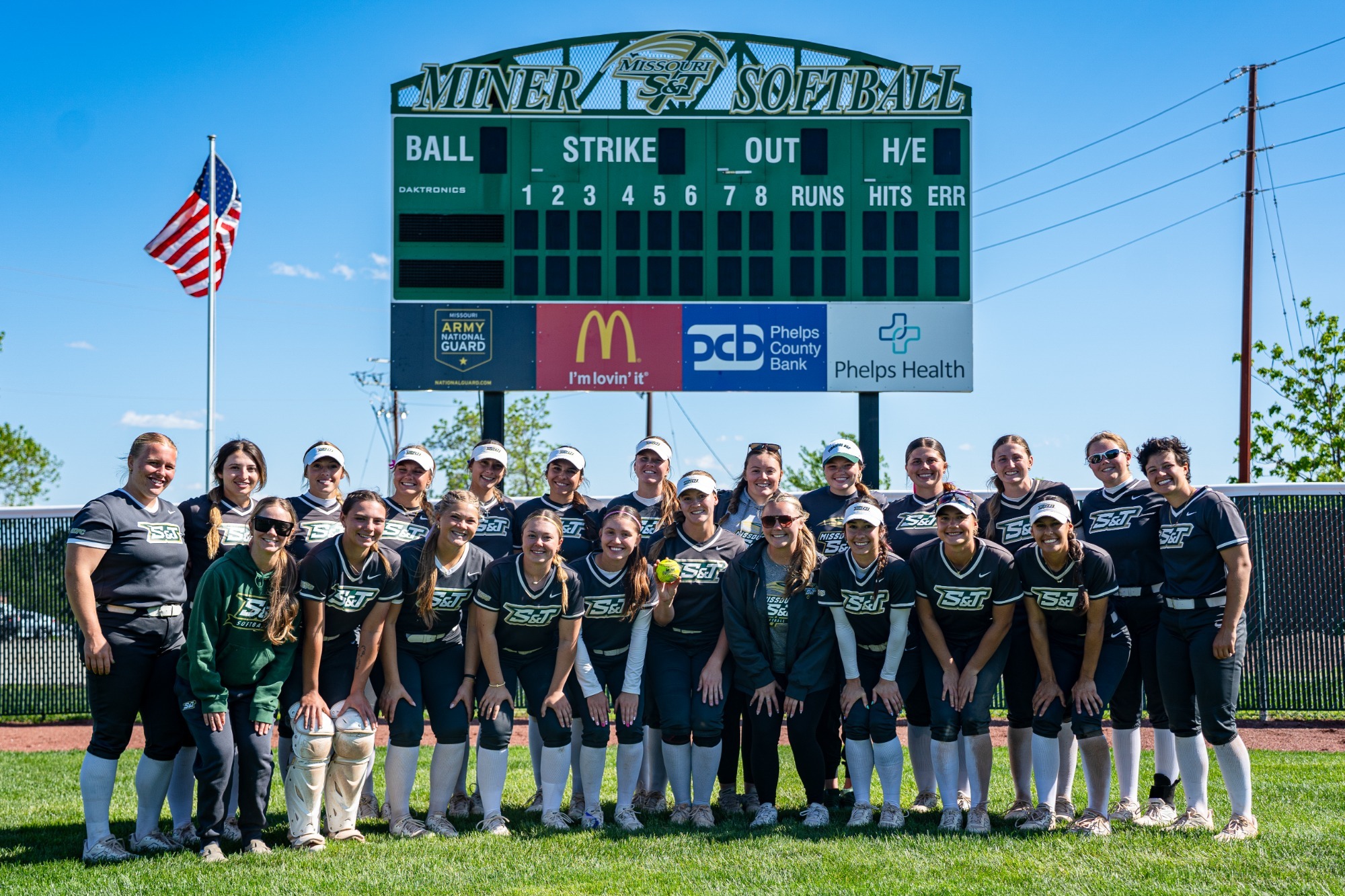 2026 Women's Softball Team Photo with team standing in the outfield under the scoreboard while wearing dark gray jerseys