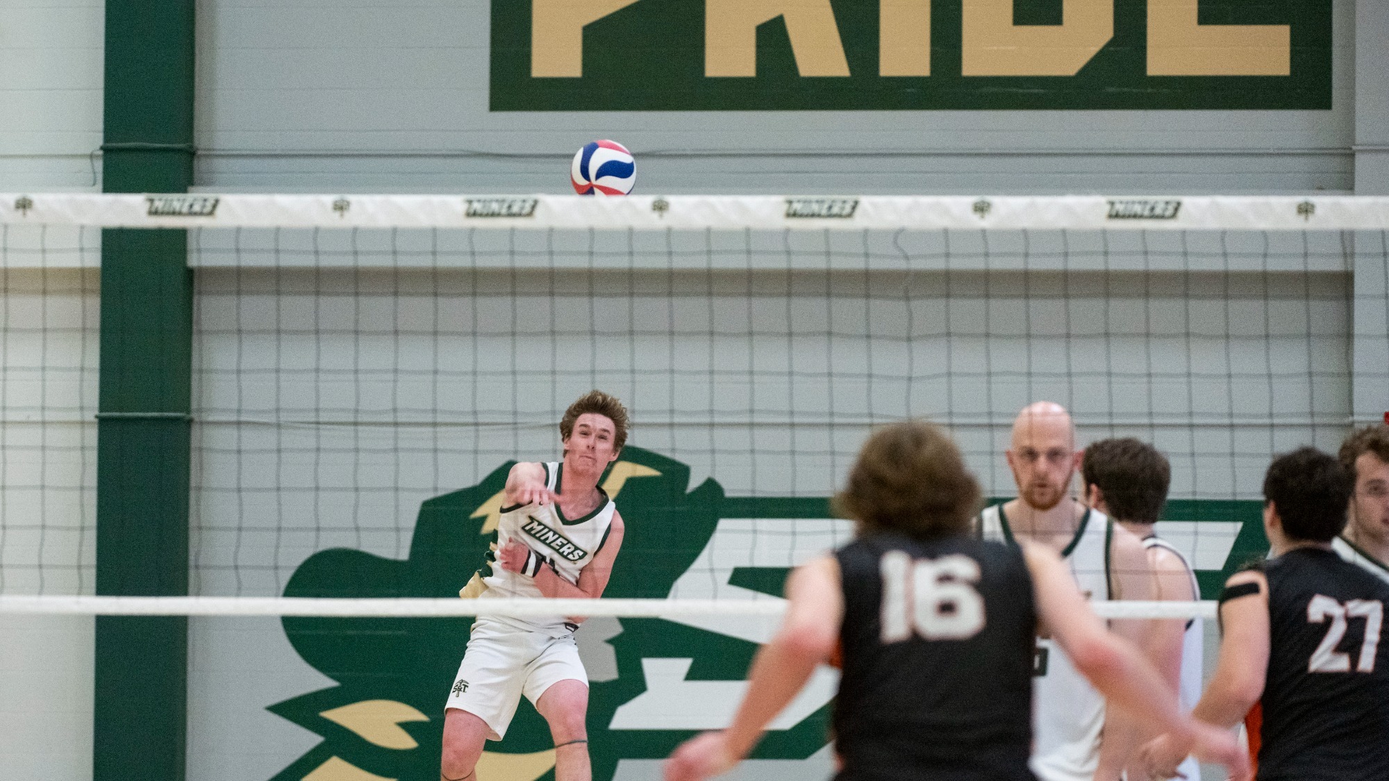 Grant Edmonds photographed through the net, leaping in the air and striking the ball while serving. He is wearing a white uniform with green highlights