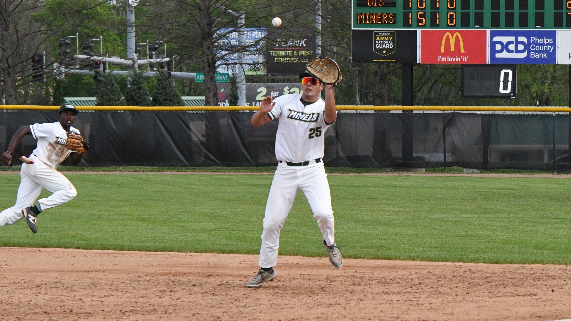 Missouri S&T baseball's Nick Hooper is photographed while leaping off the ground with his glove outstretched to catch a line drive ball headed towards him at first base. He is wearing a white uniform with green trim along with a green cap and sunglasses