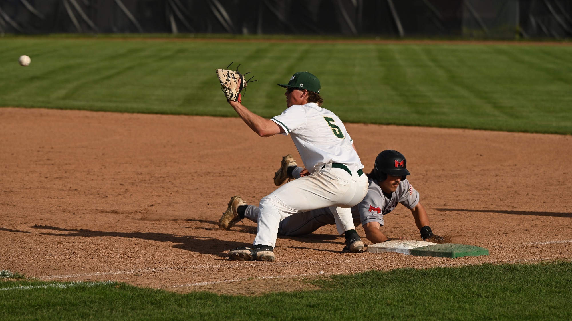 Denton Damgaard in a white uniform reaches forward with his glove to catch a throw while straddling second base as a runner in a gray uniform slides headfirst toward the bag. The ball is visible in midair approaching the fielder’s glove, and both players are focused on the play on a dirt infield with a grassy outfield in the background.