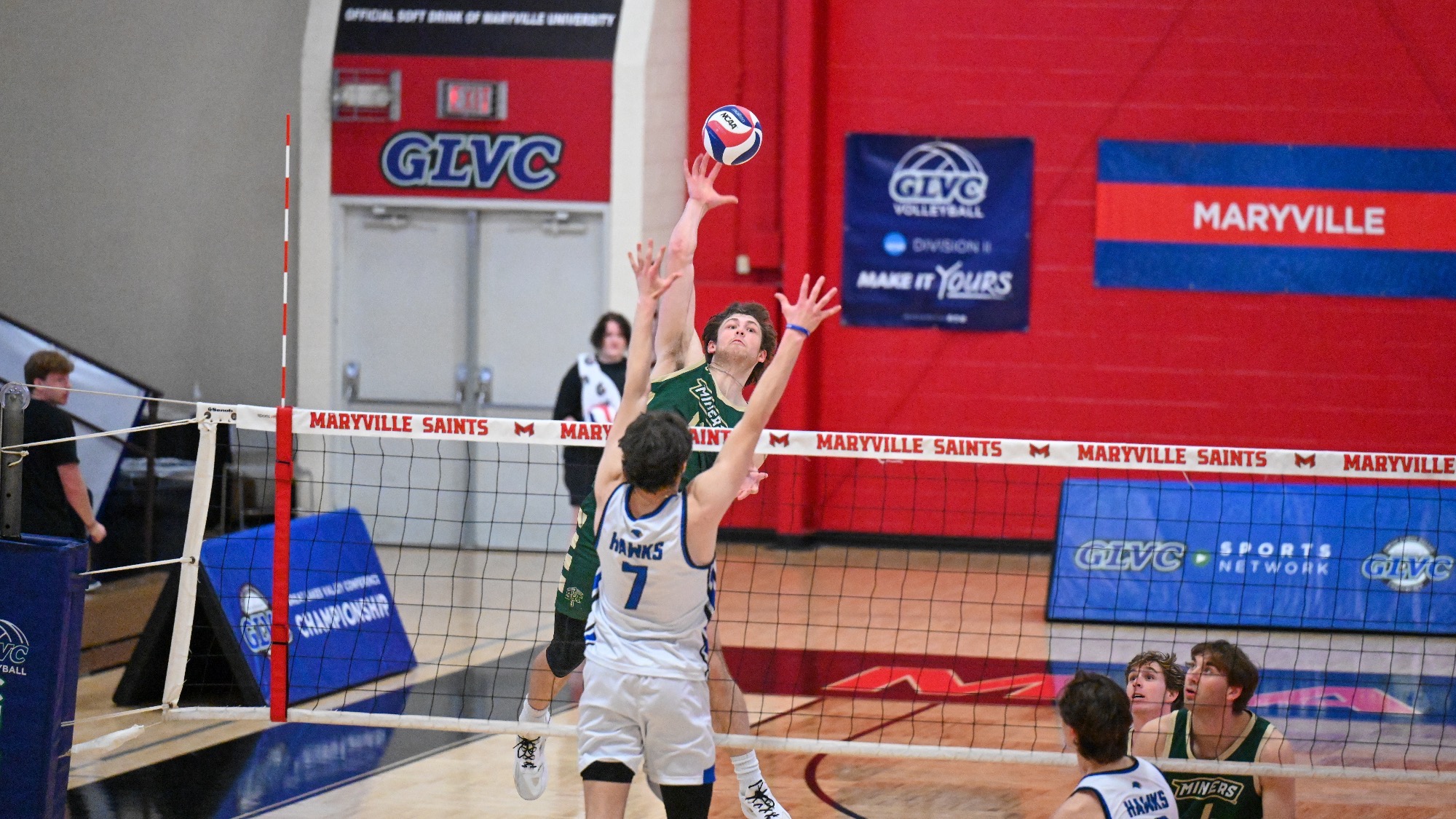 Missouri S&T’s Aaron Sallade elevates at the net to attack as a Rockhurst blocker (#7) jumps to challenge, with teammates watching from the right side.